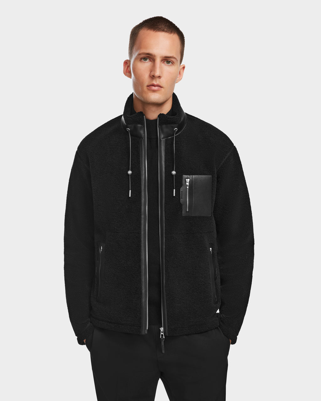 A man with short hair faces forward against a plain white background, wearing the Rudsak JOSEPH - 6524535 Black jacket featuring a high collar, drawstrings, and a zippered chest pocket.