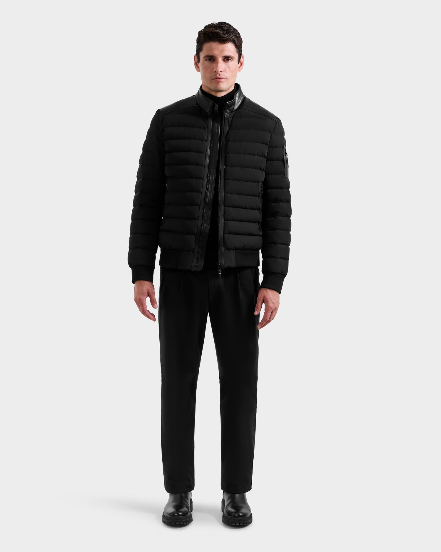 A man stands facing forward wearing the Rudsak JOSHUA - 6125722 Black down jacket with black pants and shoes against a plain white background.