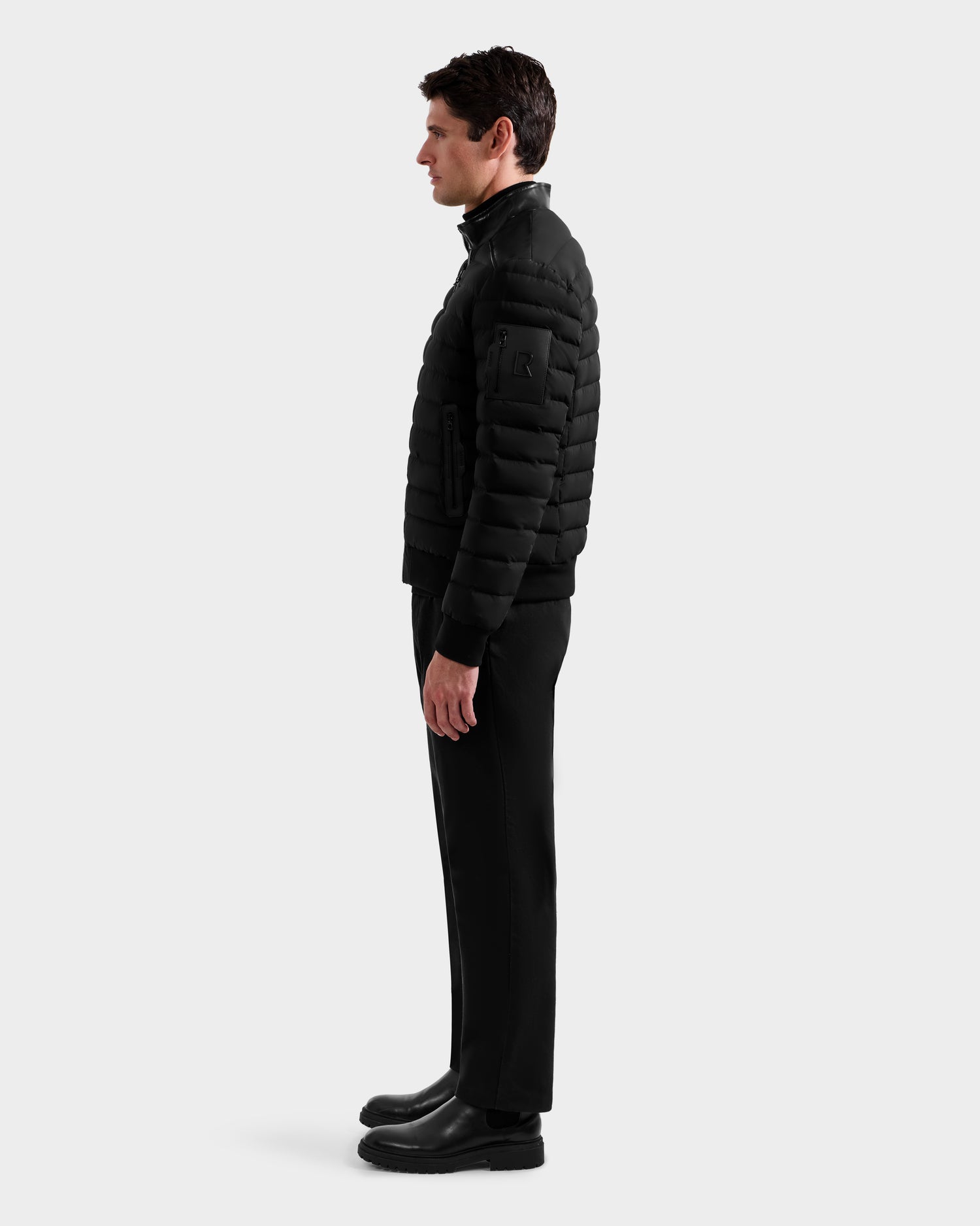 A man stands in profile against a plain white background, wearing the Rudsak JOSHUA - 6125722 Black men's down jacket, styled with black pants and boots.