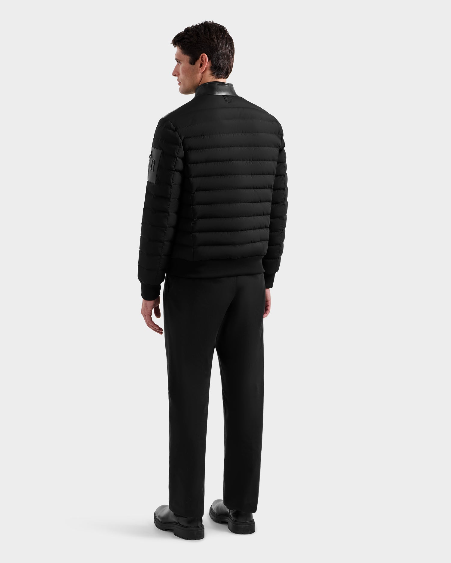 A man stands facing away in a Rudsak JOSHUA - 6125722 Black men’s down jacket, paired with black pants and shoes against a plain light-colored background.