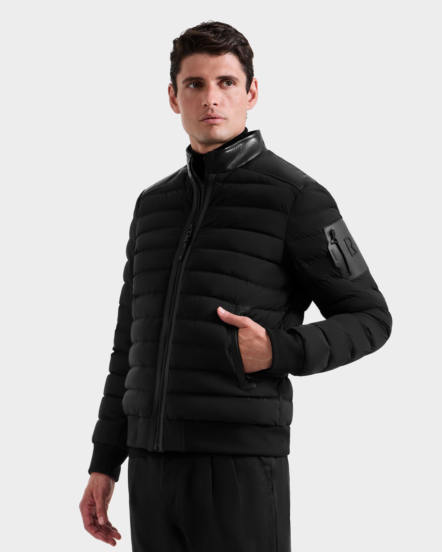 A man wears the Rudsak JOSHUA - 6125722 Black jacket, featuring a quilted design and a zippered pocket on the upper left sleeve, standing with one hand in his pocket against a plain light background.