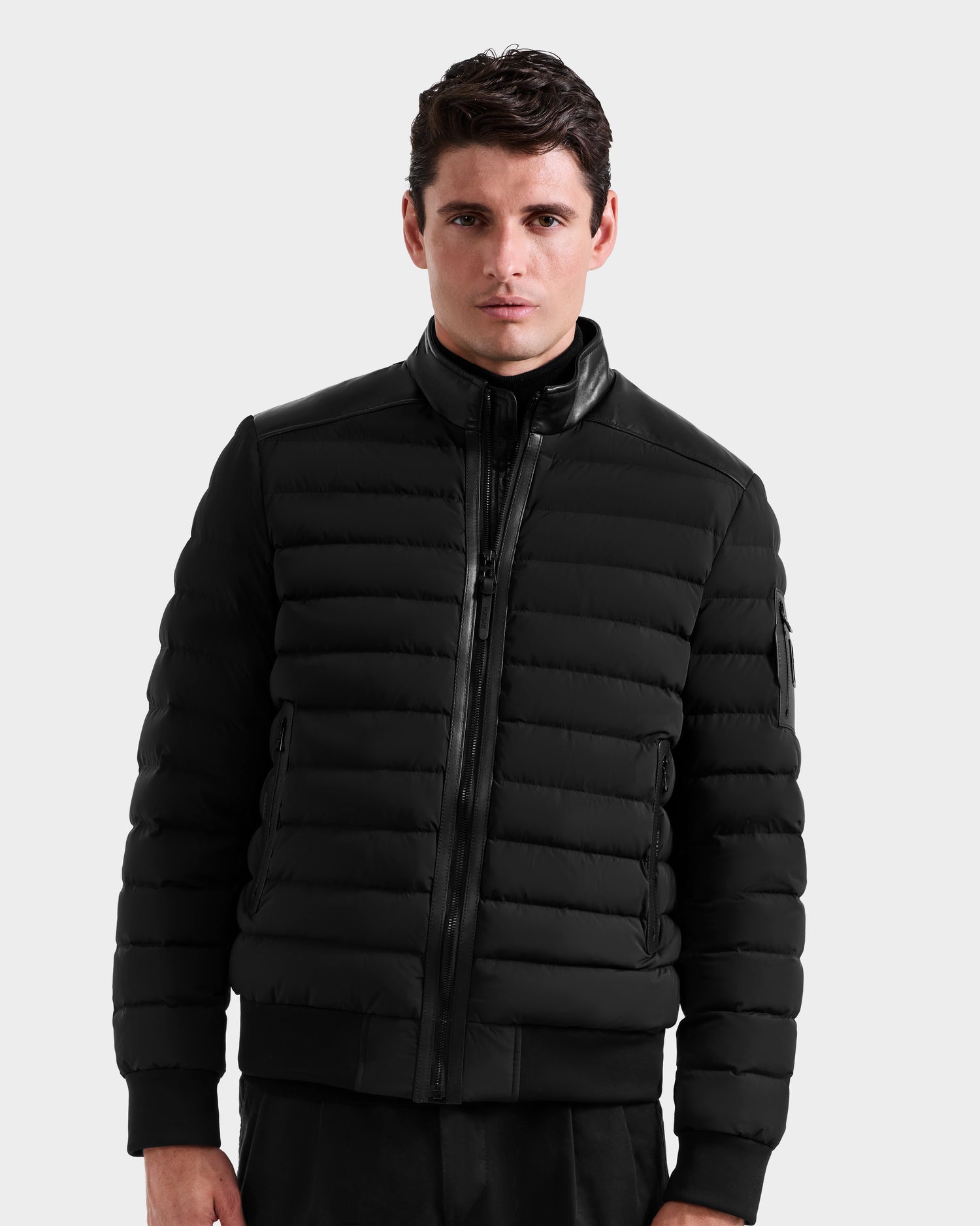 A man wears the Rudsak JOSHUA - 6125722 Black quilted down jacket with a zip-up front, high collar, and side pockets, standing against a plain light gray background.