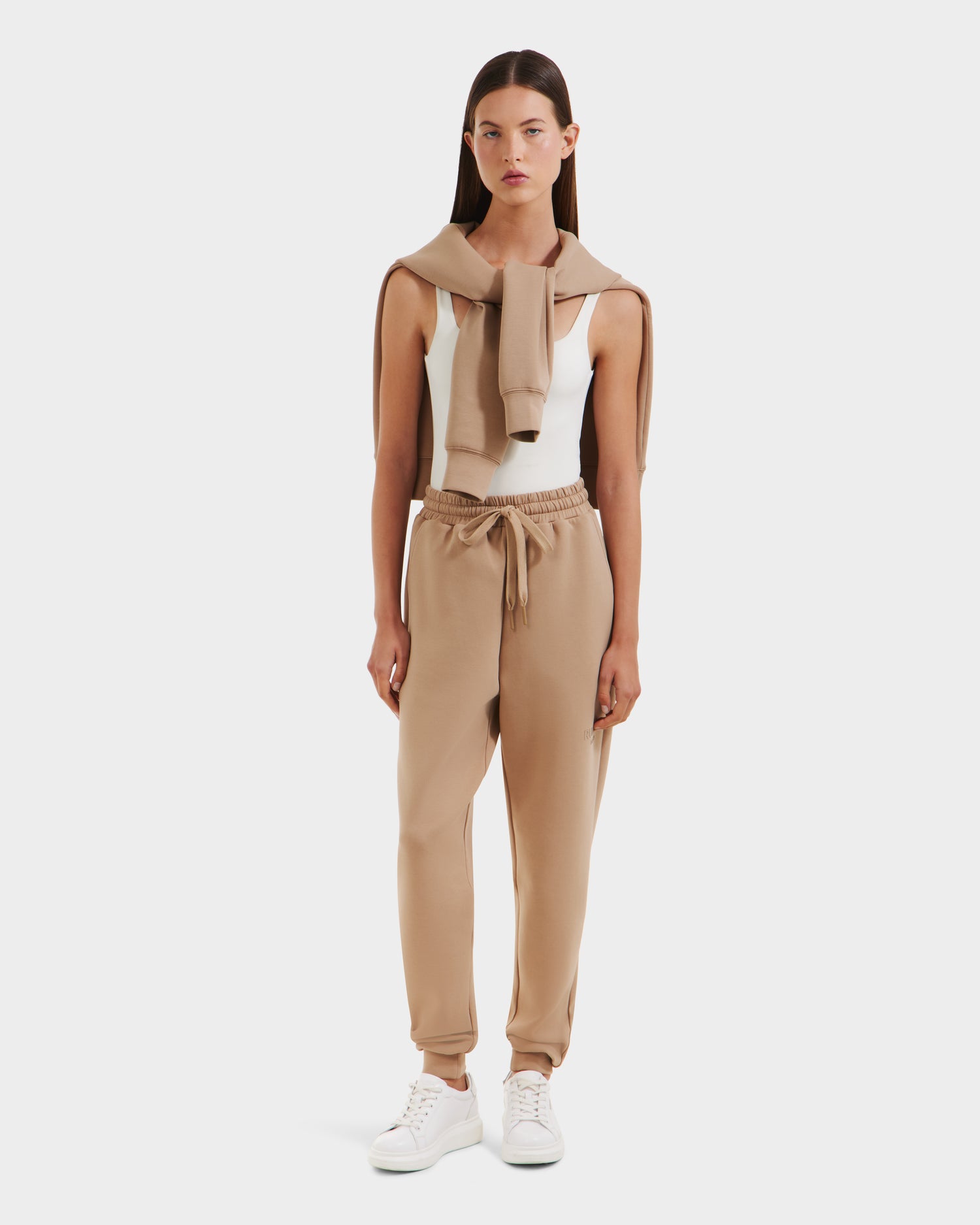 A woman faces forward against a white background, wearing the Rudsak JOSS - 9524009 Almond W premium cotton joggers in tan, a matching sweater over her shoulders, a white tank top, and white sneakers.