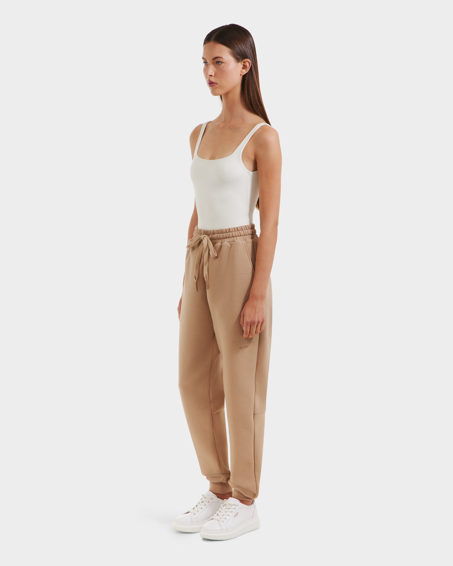 A woman stands against a white background, wearing Rudsaks JOSS - 9524009 Almond W beige joggers with a drawstring waist, paired with a white tank top and sneakers. She is facing forward, arms by her sides.
