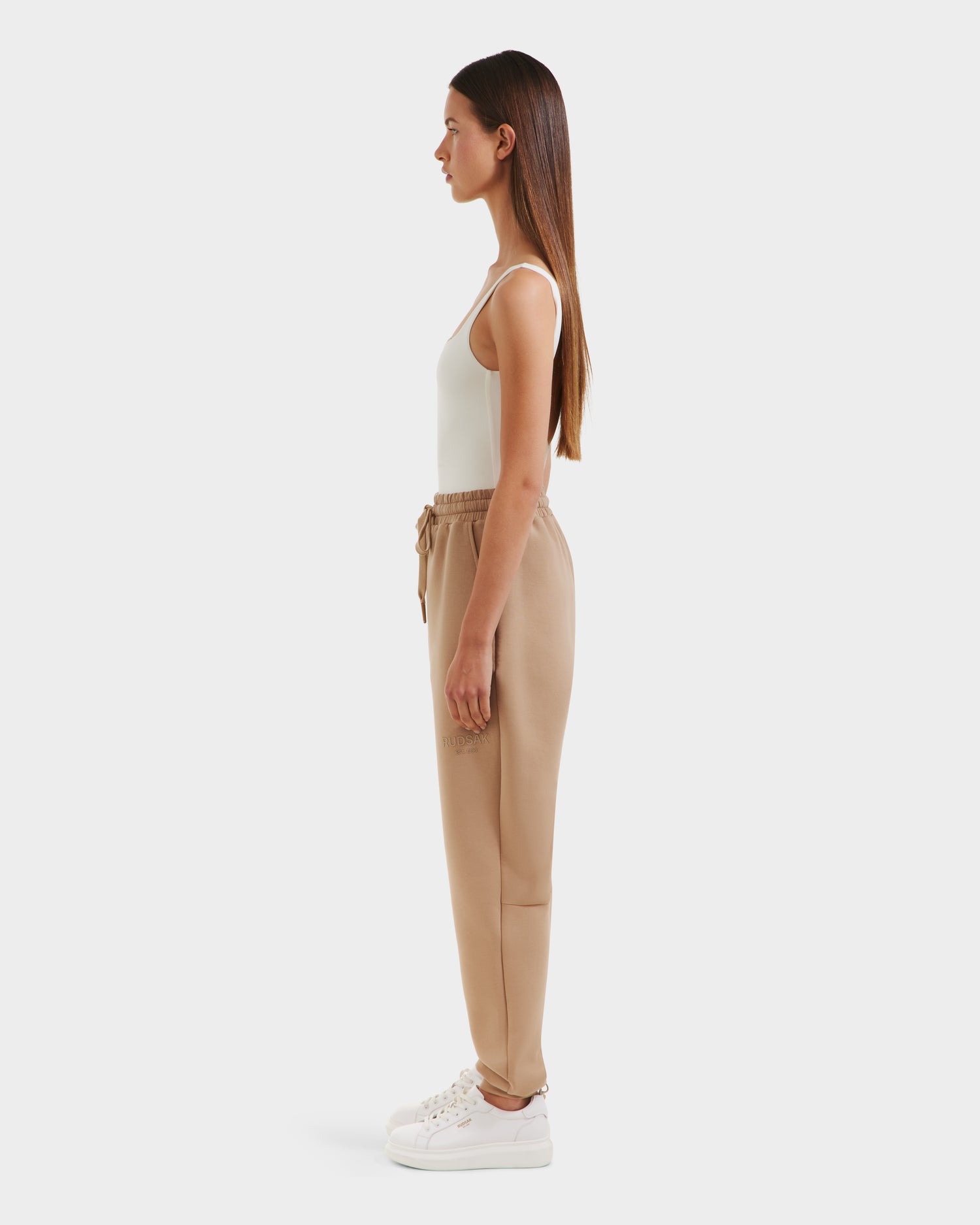 A woman stands in profile facing left, wearing Rudsaks JOSS - 9524009 Almond W white platform sneakers, a white tank top, and premium cotton joggers with a drawstring. She has long straight brown hair and a neutral expression against a plain white background.