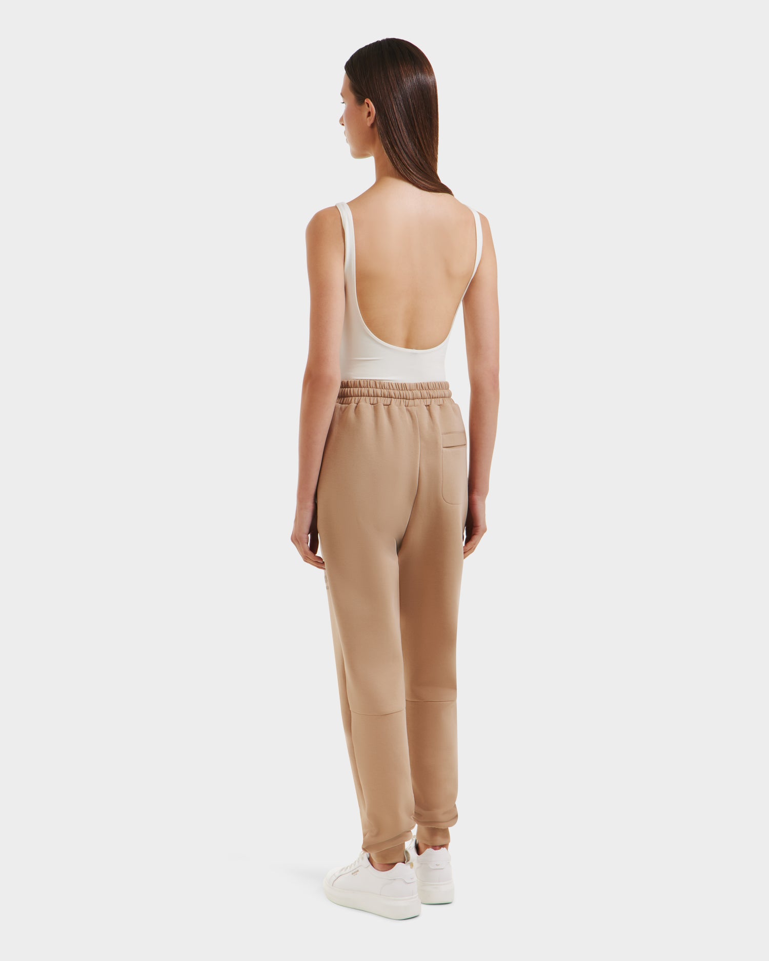 A woman faces away, wearing Rudsak’s JOSS - 9524009 Almond W sleeveless, low-back beige top, light brown joggers, and white sneakers against a plain white background.