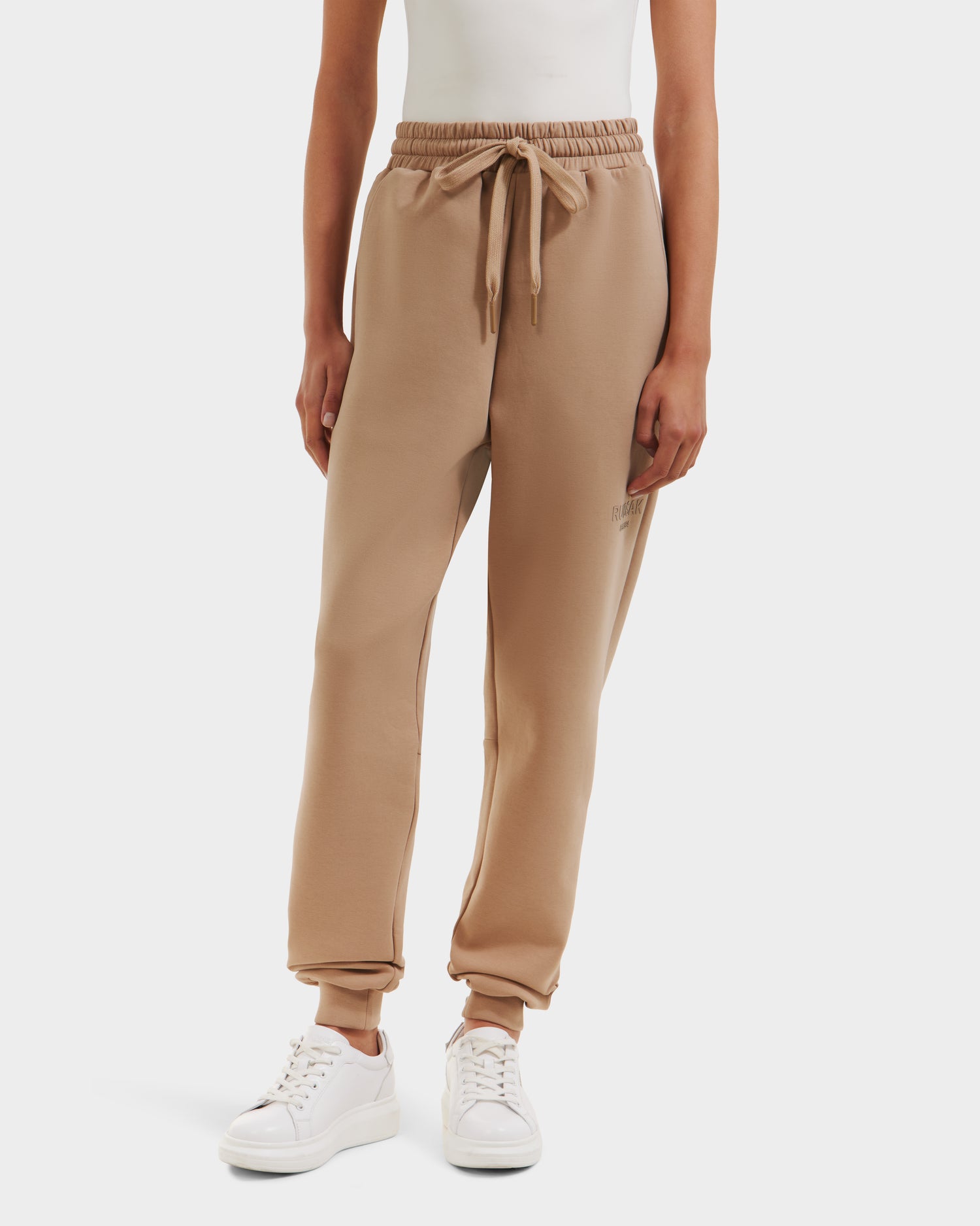 A person wearing Rudsak JOSS - 9524009 Almond W joggers with an elastic waistband and drawstring, paired with a white sleeveless top and white sneakers, stands against a plain white background.