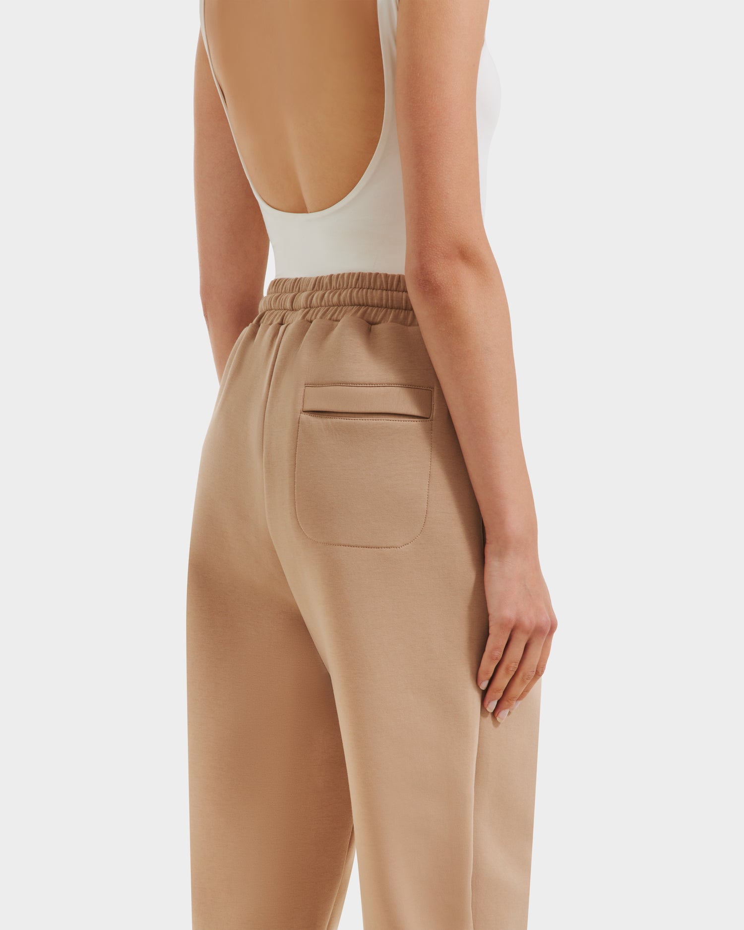 A person is shown from the back wearing Rudsak’s JOSS - 9524009 Almond W tan high-waisted joggers and a white sleeveless low-back top, with one arm resting at their side.