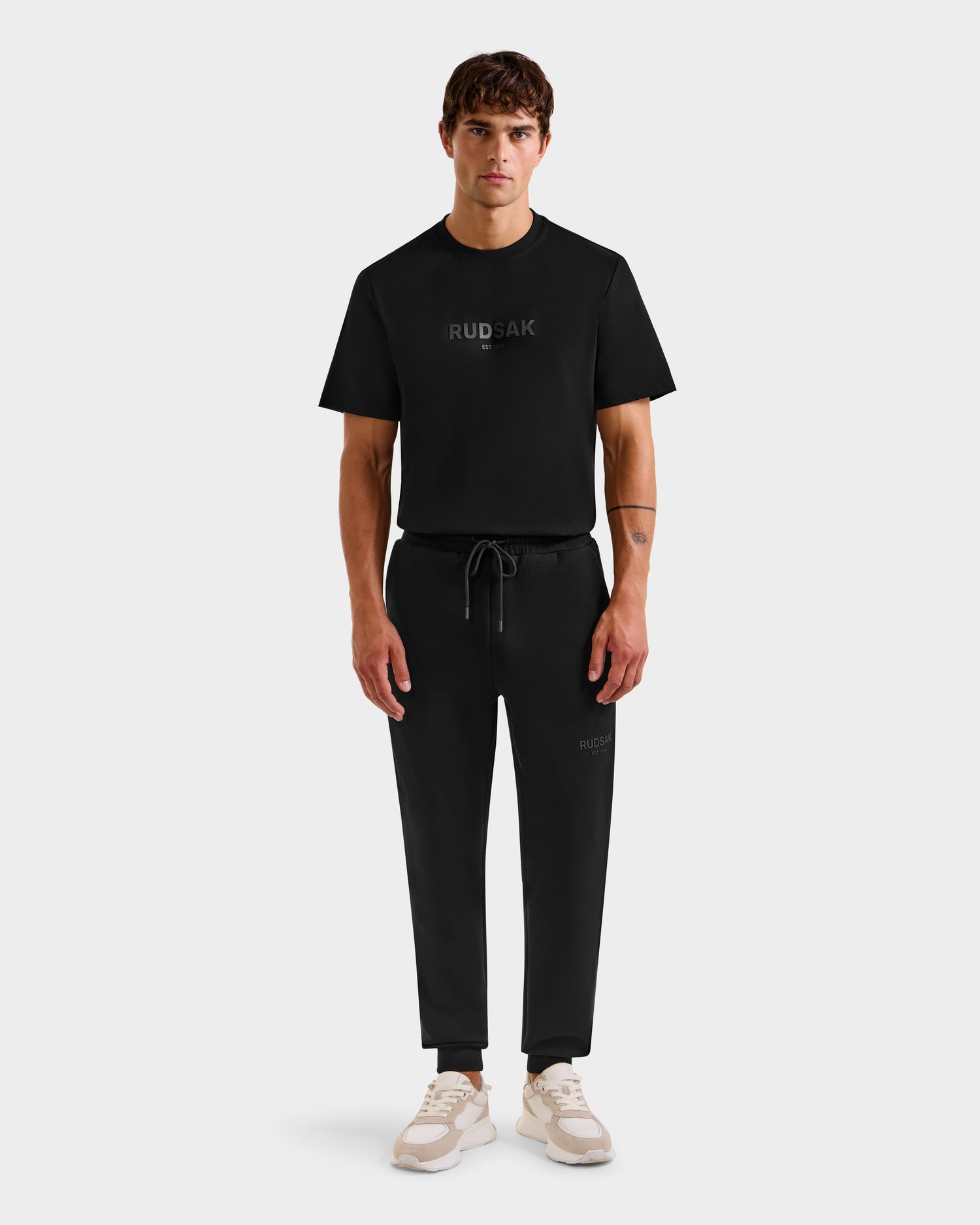 A man faces forward wearing the Rudsak JOSS - 9524009 Black M t-shirt and stylish joggers with branding, styled with white sneakers against a plain light gray background.