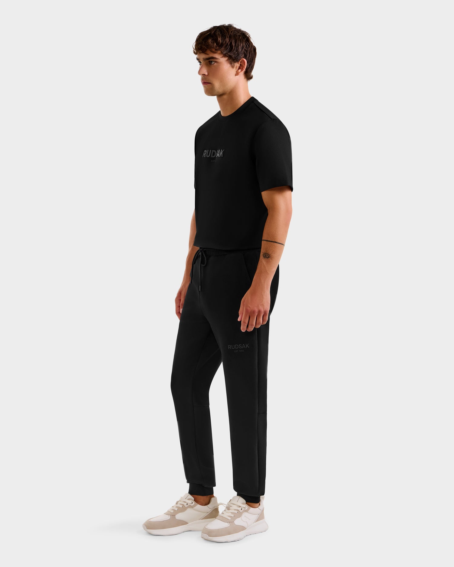 A man stands against a plain background wearing the Rudsak JOSS - 9524009 Black M T-shirt. He is looking slightly to the side with his arms relaxed by his sides.