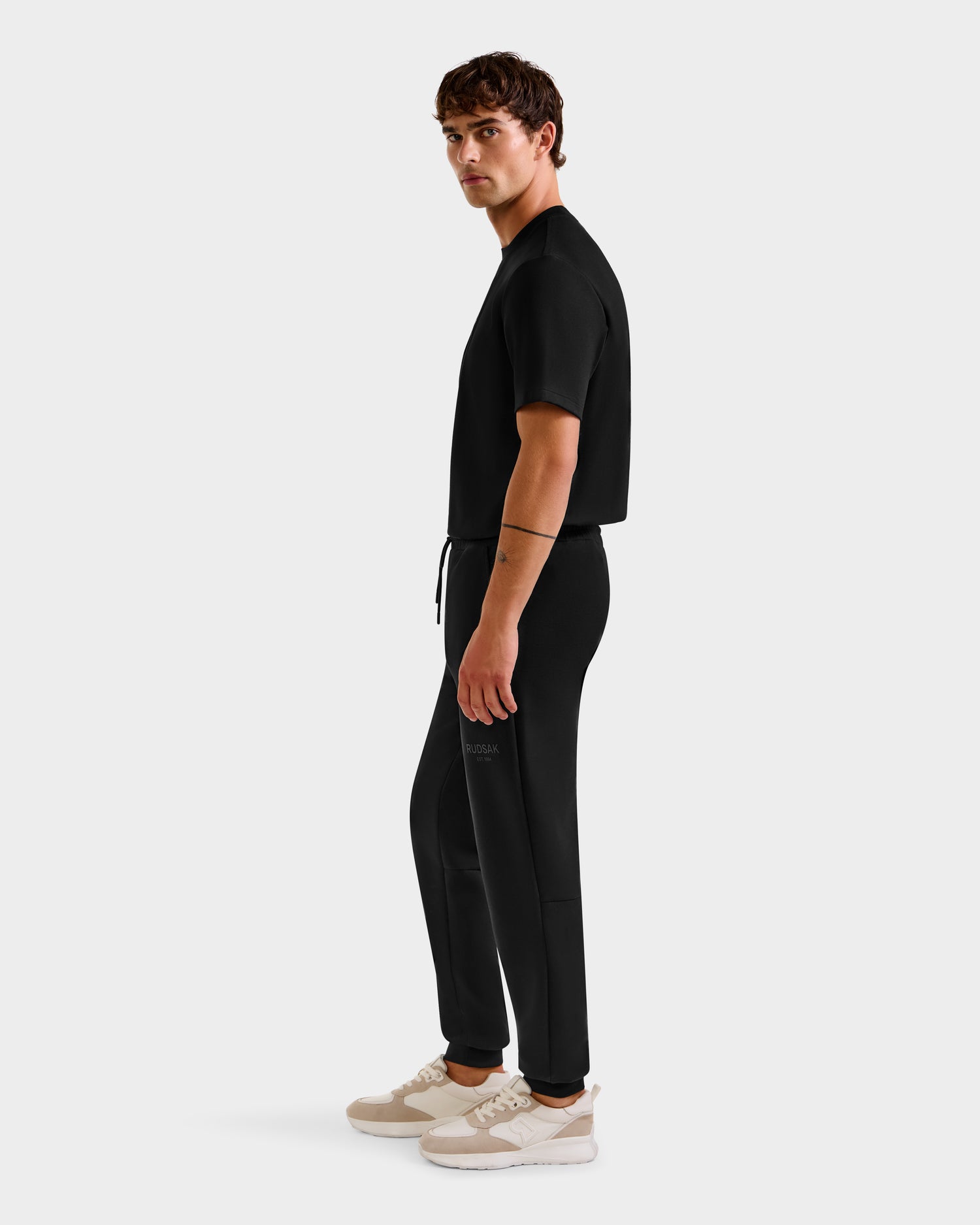 A person stands sideways against a plain white background, wearing the Rudsak JOSS - 9524009 Black M short-sleeve shirt paired with premium cotton joggers and white sneakers.