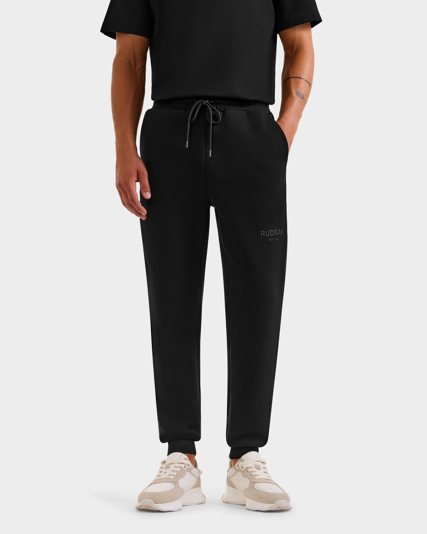A person wears Rudsak's JOSS joggers in black (M), featuring a drawstring waist and RUDSAK branding on the left thigh, paired with a black tee and beige sneakers. The background is plain white.