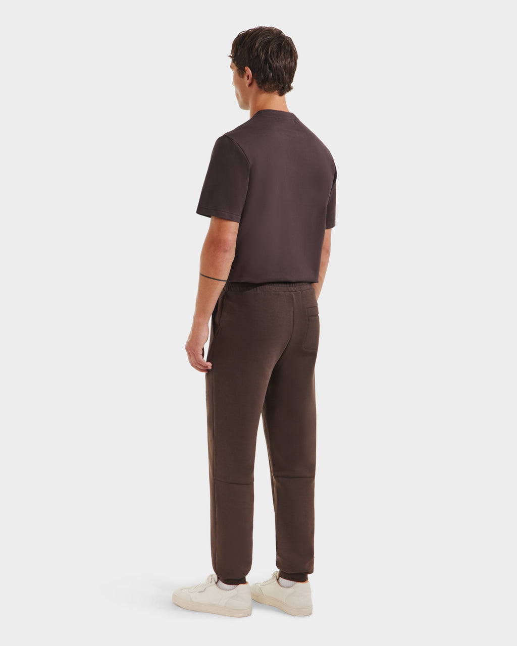 A person faces away, wearing the Rudsak JOSS - 9524009 Espresso M short-sleeve shirt with stylish joggers and white sneakers, set against a plain white background.