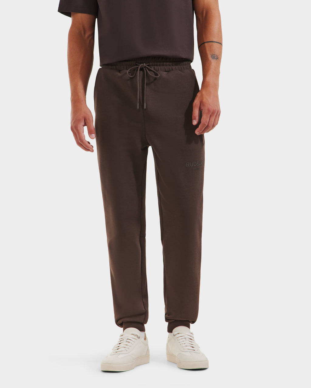 A person models the Rudsak JOSS - 9524009 Espresso M jogger sweatpants, styled with a dark t-shirt and white sneakers. The photo, cropped at the shoulders against a plain white background, highlights the premium cotton blend pants.