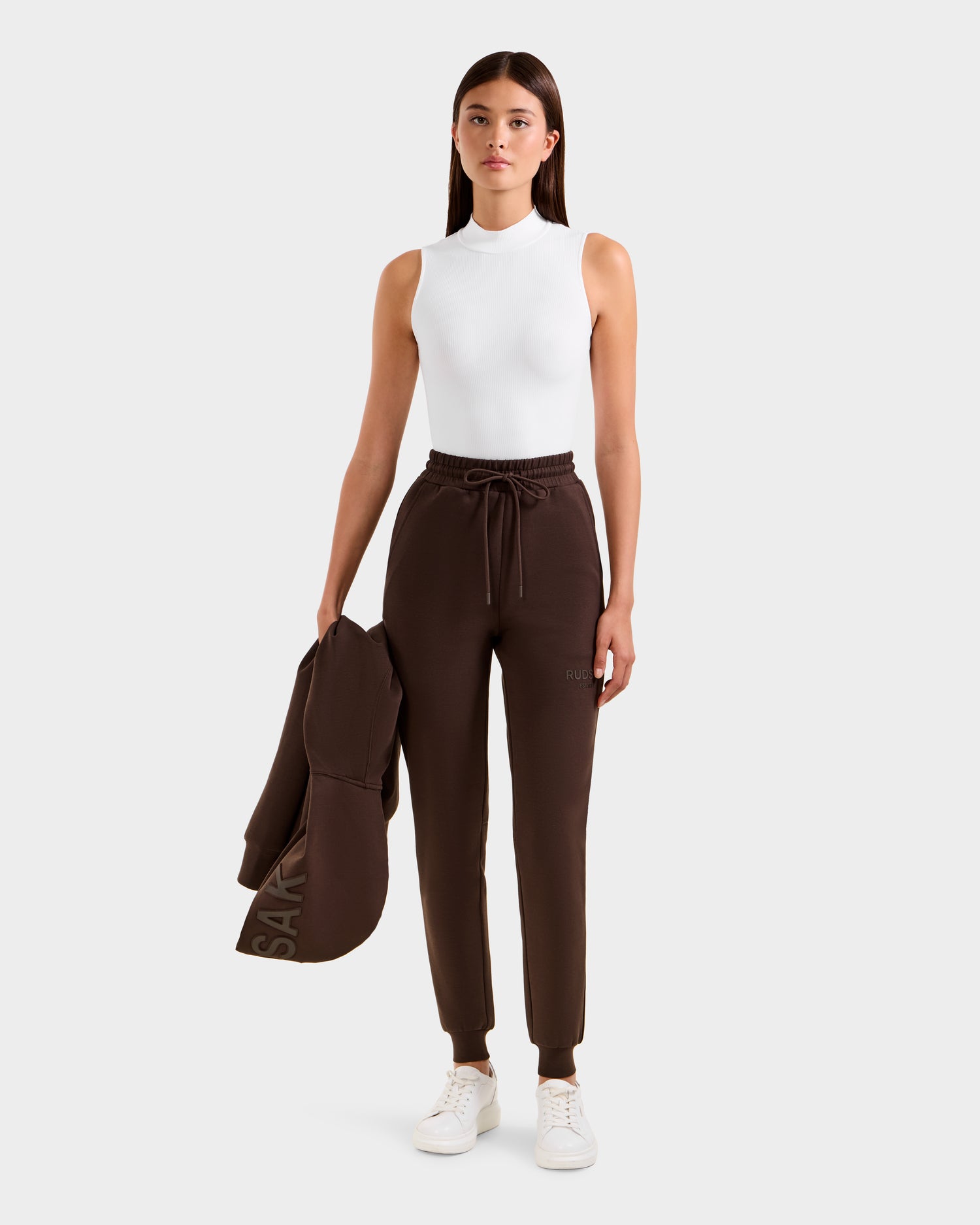A woman faces forward against a plain light background, wearing the Rudsak JOSS - 9524009 Espresso W joggers and holding the matching brown jacket in her left hand, paired with a sleeveless white mock-neck top and white sneakers.