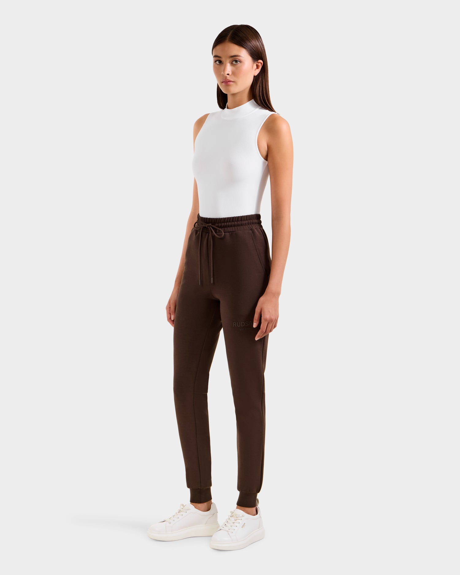 A woman faces forward wearing Rudsak’s JOSS - 9524009 Espresso W modern dark brown jogger pants with a drawstring, paired with a sleeveless white top and white sneakers against a plain light background.