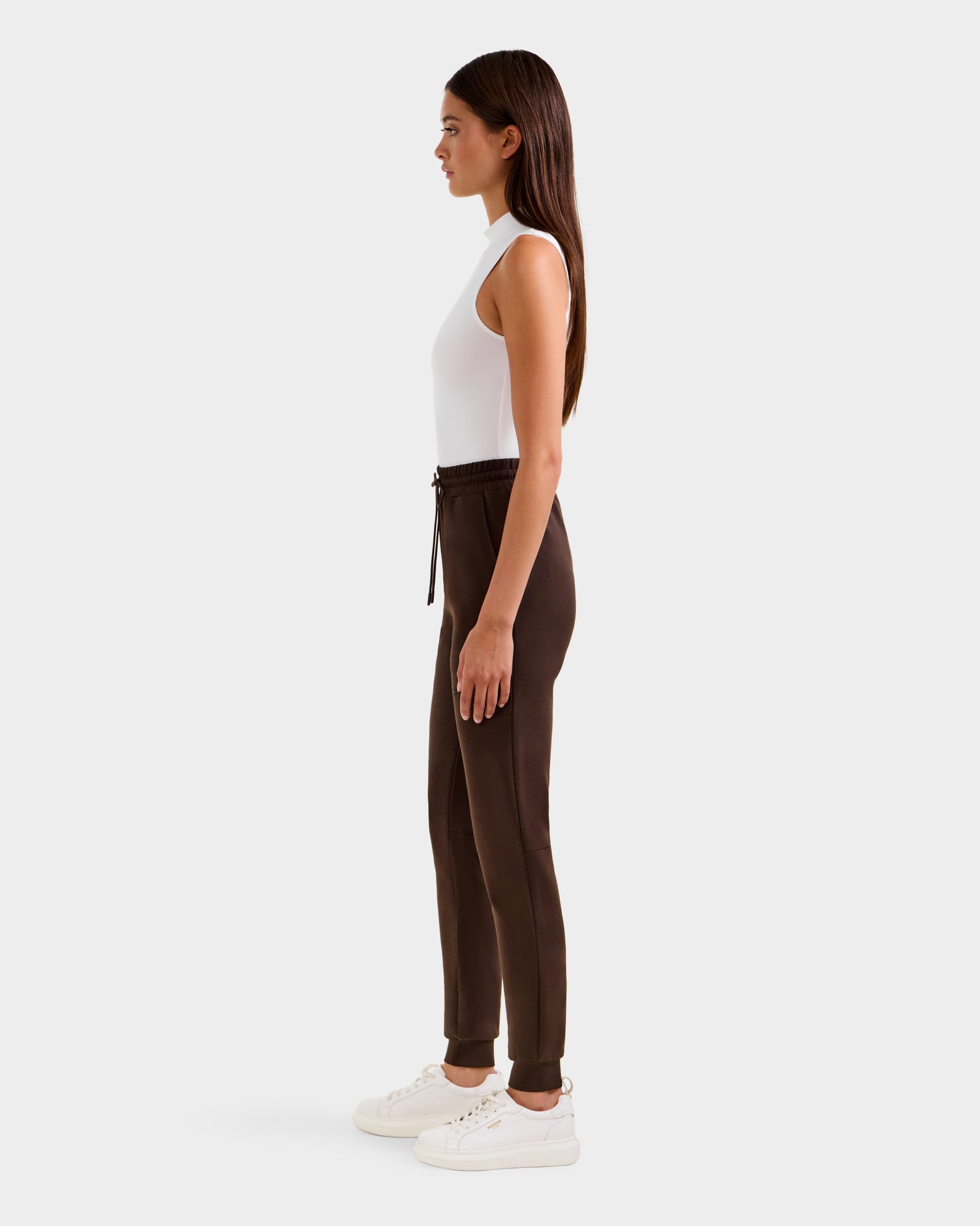 A woman in profile against a light background wears the Rudsak JOSS - 9524009 Espresso W joggers with a drawstring waist, a sleeveless white top, and white sneakers. Her straight long hair flows as she faces left.