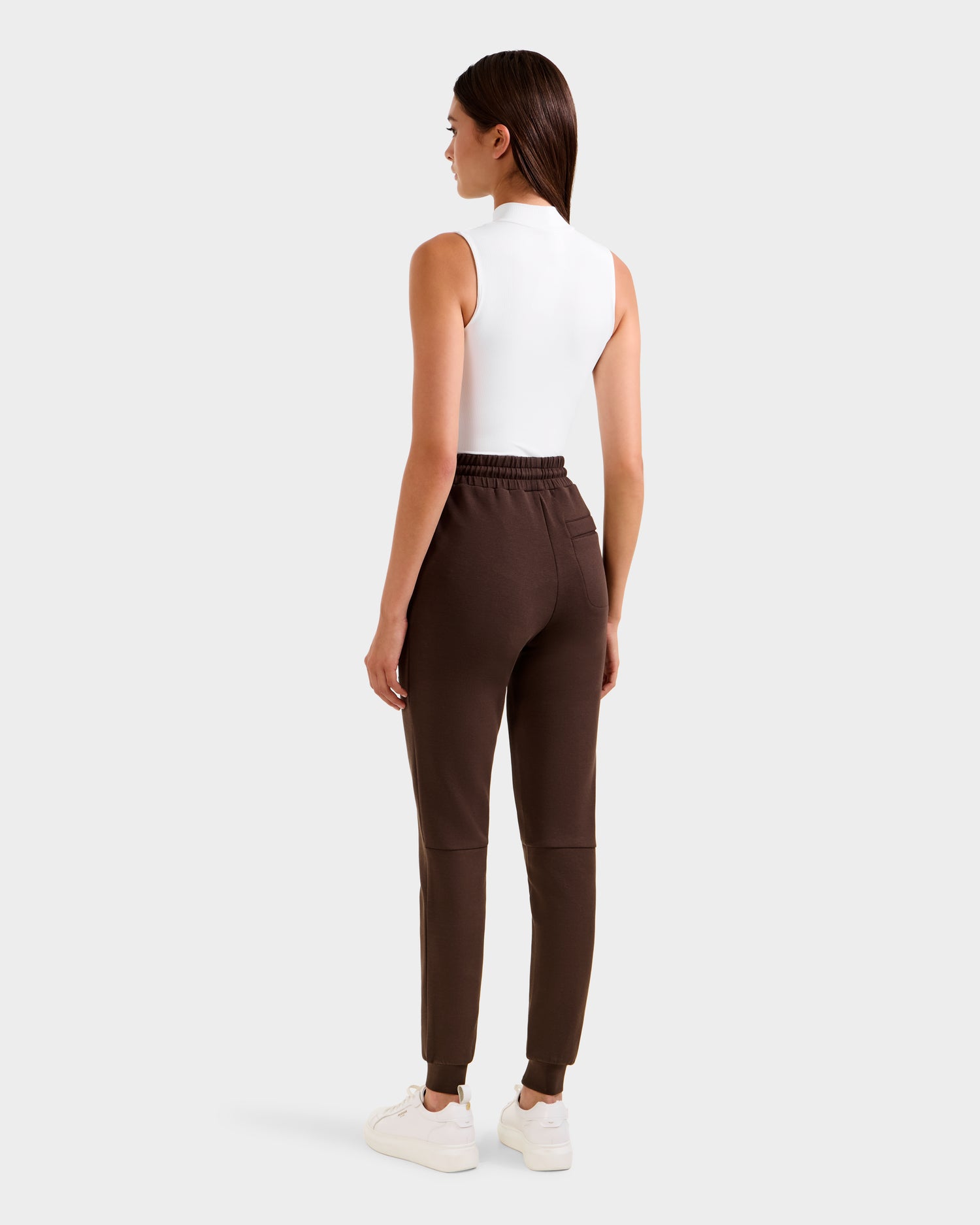 A woman stands facing away, wearing Rudsak's JOSS - 9524009 Espresso W sleeveless white top and high-waisted premium cotton joggers with white sneakers, set against a plain light gray background.