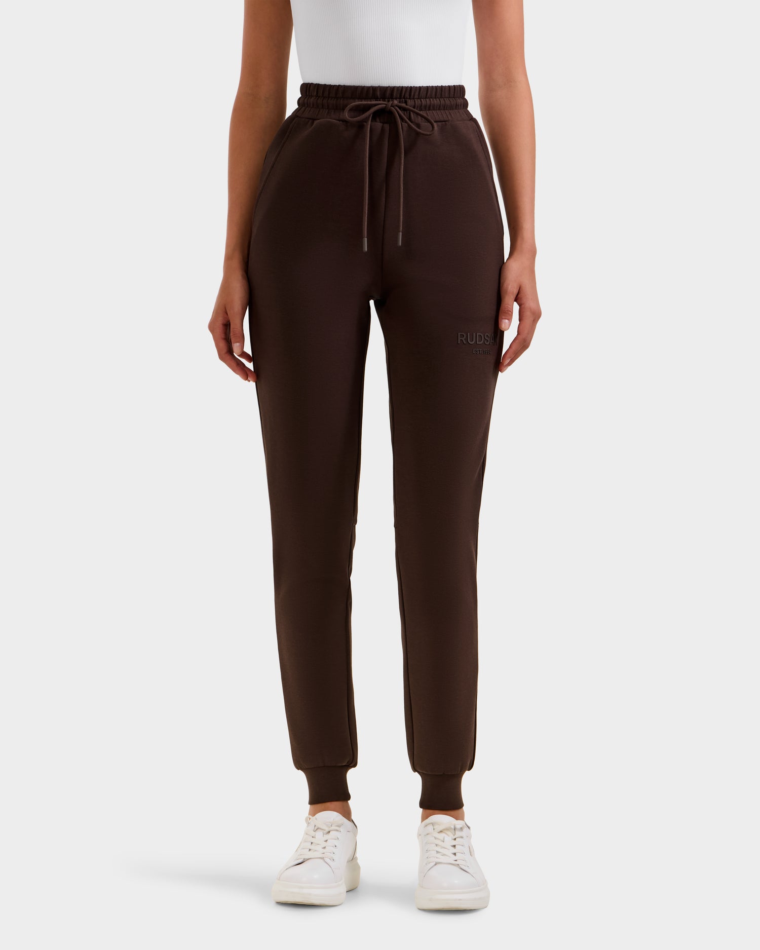 A person wears Rudsak’s JOSS - 9524009 Espresso W premium cotton joggers with a drawstring waist, a white sleeveless top, and white sneakers, standing against a plain light background.