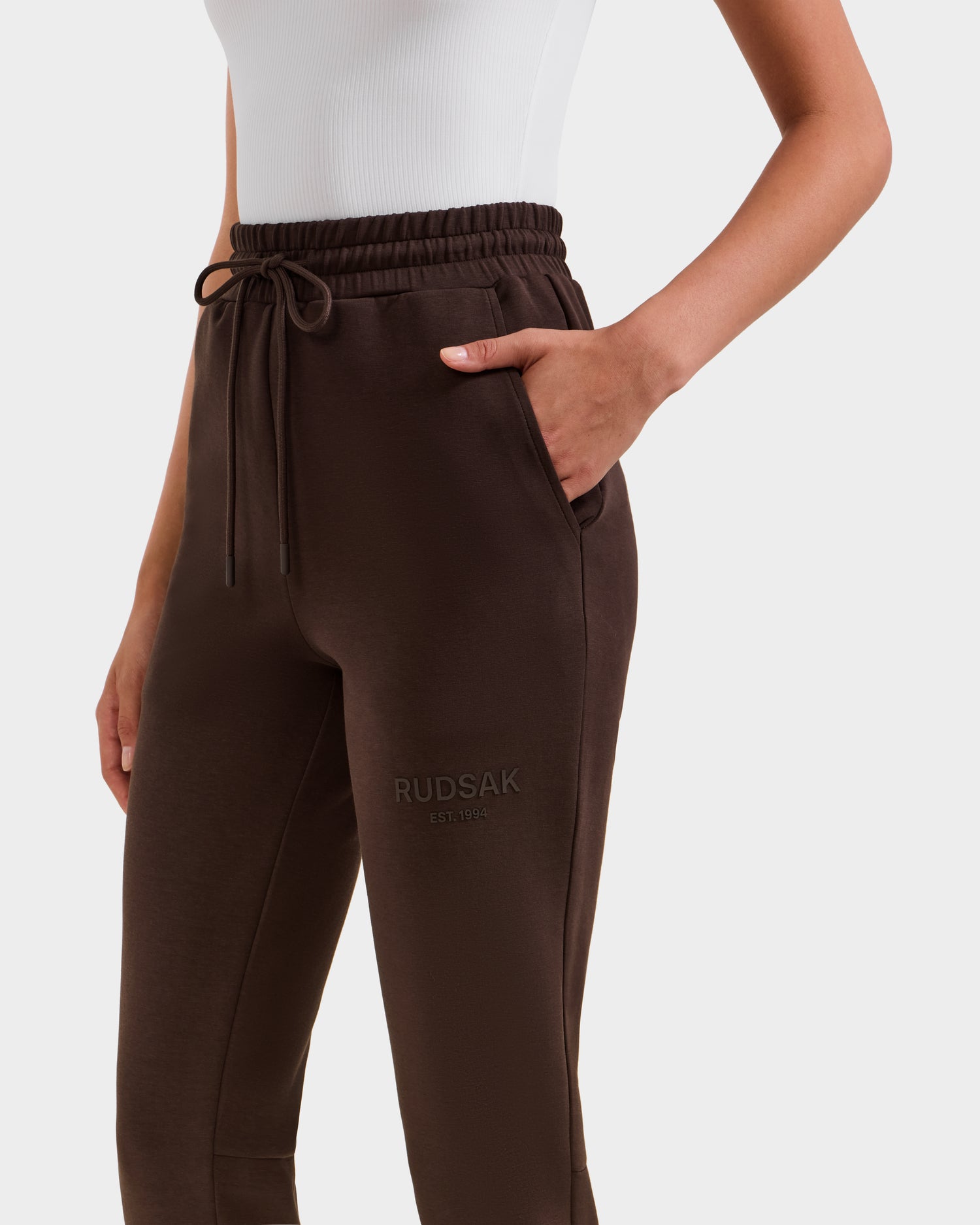 A person wears a white sleeveless top and Rudsak JOSS - 9524009 Espresso W premium cotton joggers in dark brown with a drawstring waist and RUDSAK embossed on the upper thigh, one hand in their pocket.
