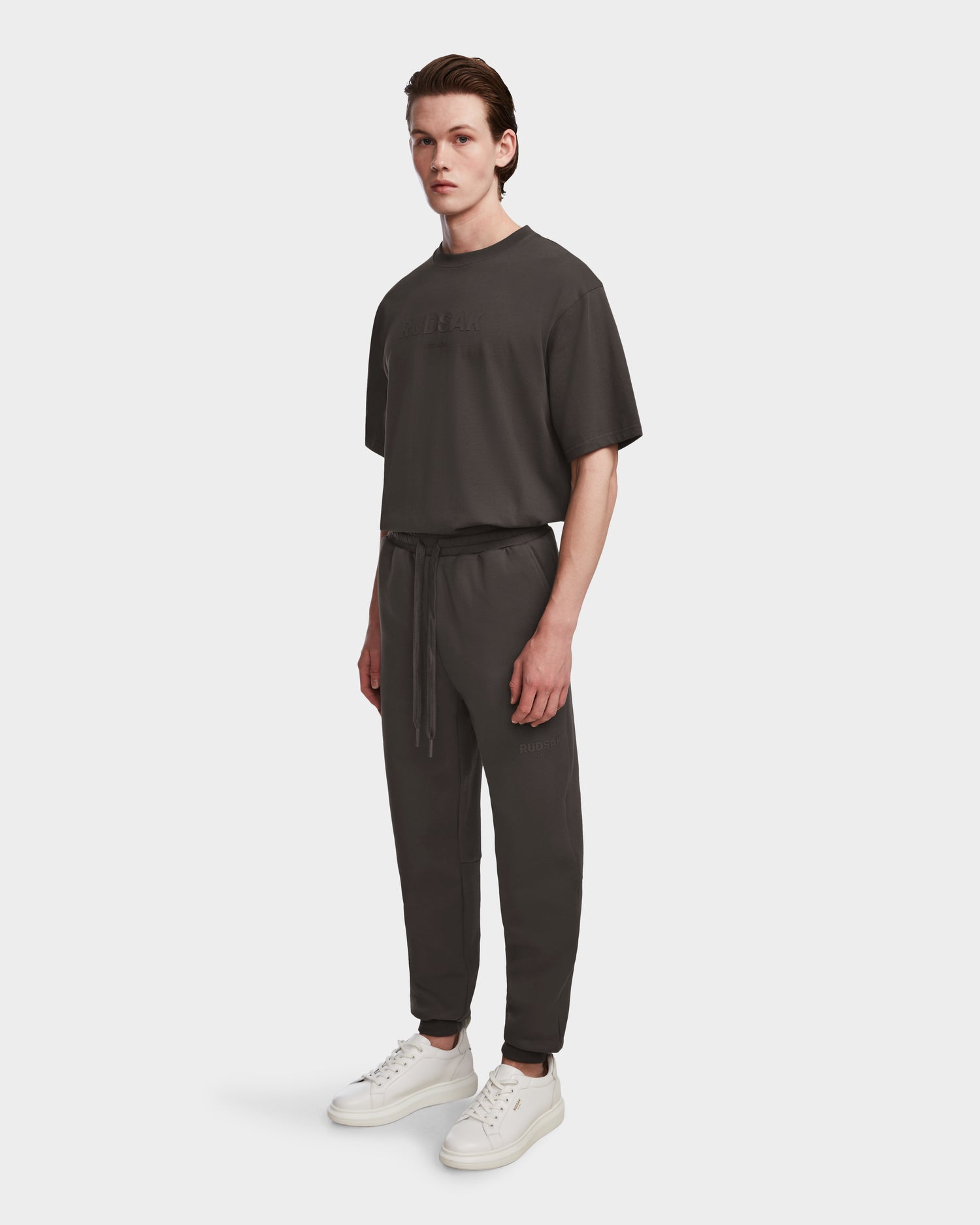A young man with straight brown hair and a neutral expression wears the Rudsak JOSS - 9524009 Grey M T-shirt against a plain white background, paired with premium cotton joggers and white sneakers.