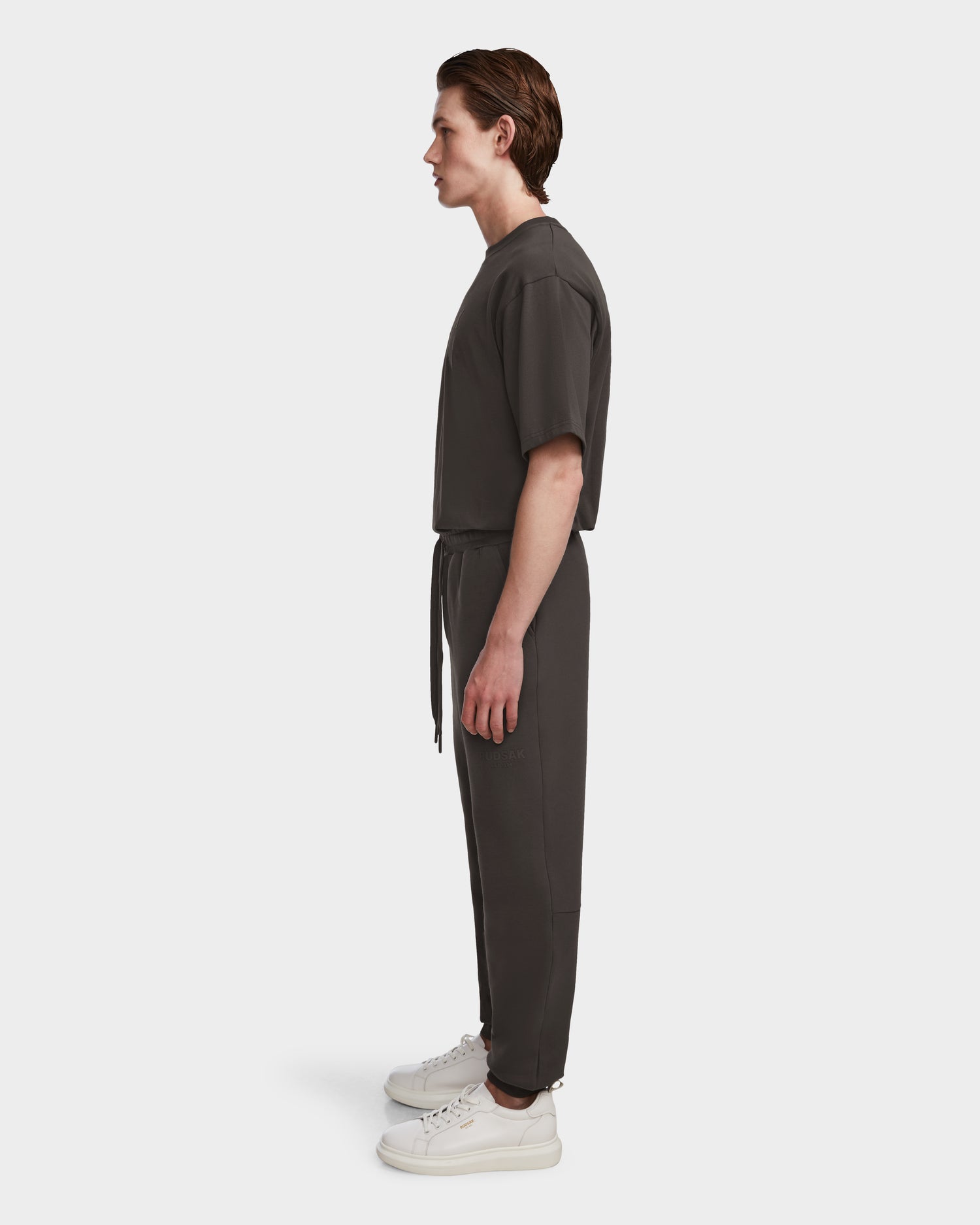 A person stands in profile against a white background, wearing the Rudsak JOSS - 9524009 Grey M joggers with a dark short-sleeve top and white sneakers.