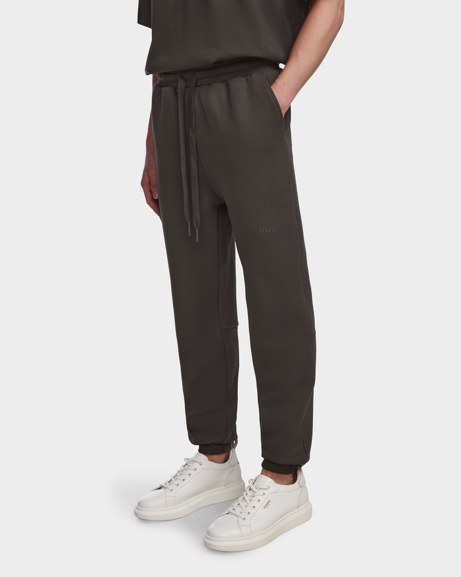 A person wears Rudsak’s JOSS - 9524009 Grey M unisex premium cotton joggers with a drawstring waist and white sneakers, standing against a plain white background—blending comfort and style for all.