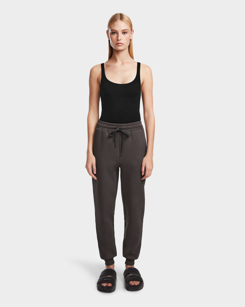 A woman stands facing forward wearing Rudsak JOSS - 9524009 Grey W premium cotton jogger pants, a black tank top, and black fuzzy slide sandals against a plain white background.