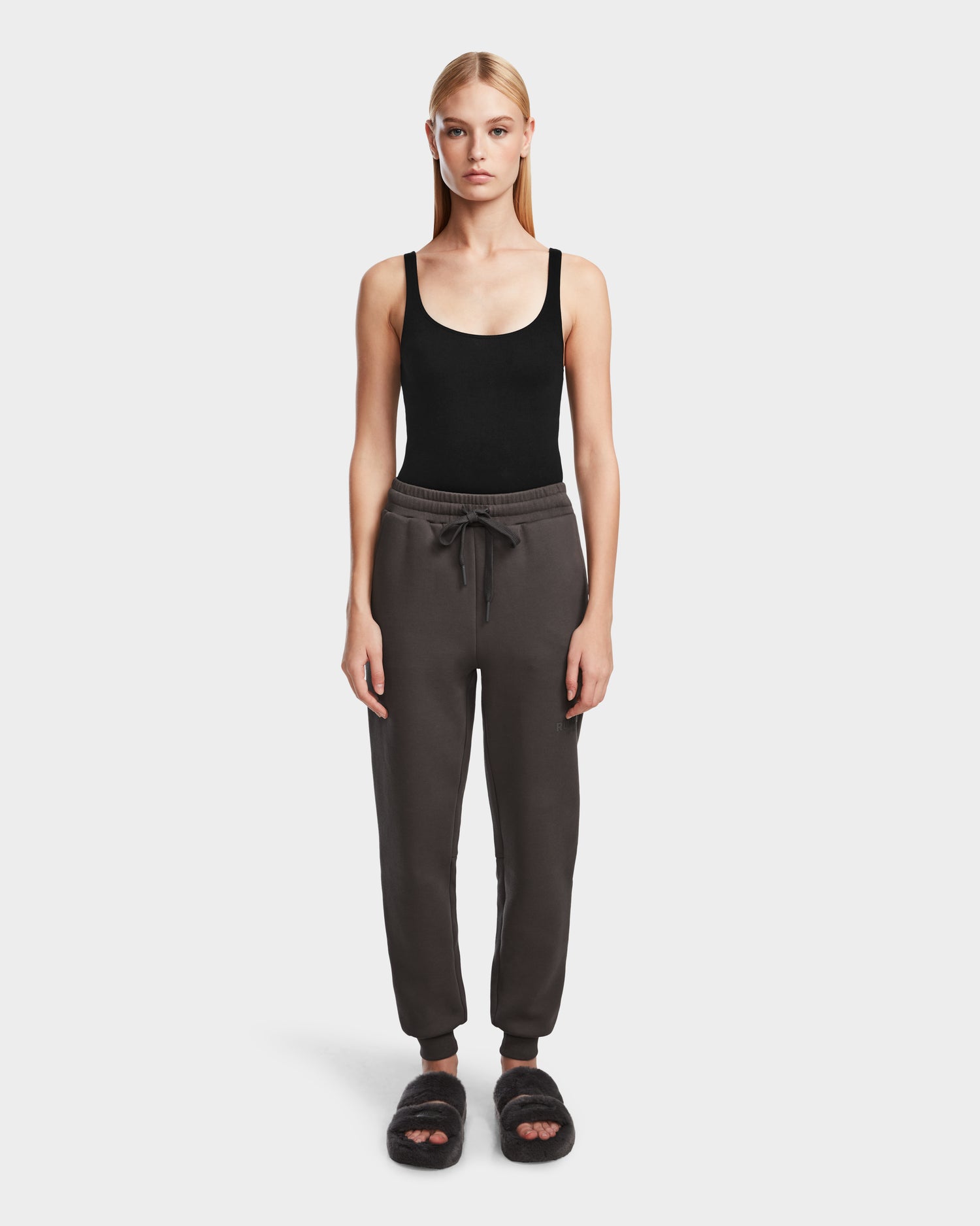 A woman stands facing forward wearing Rudsak JOSS - 9524009 Grey W premium cotton jogger pants, a black tank top, and black fuzzy slide sandals against a plain white background.