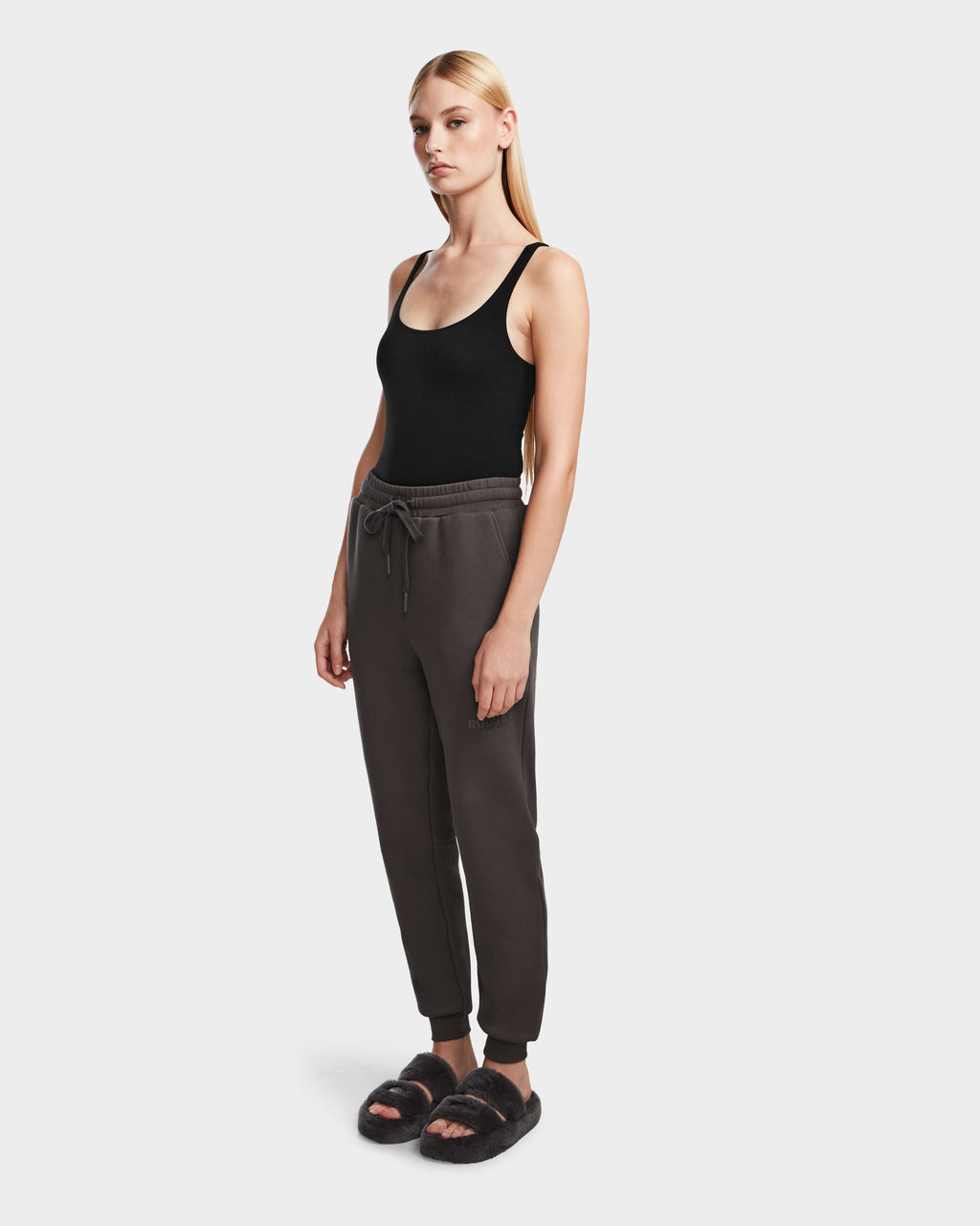 A woman with long blonde hair wears Rudsaks JOSS - 9524009 Grey W jogger pants, a black tank top, and black fuzzy slippers, standing against a plain white background.