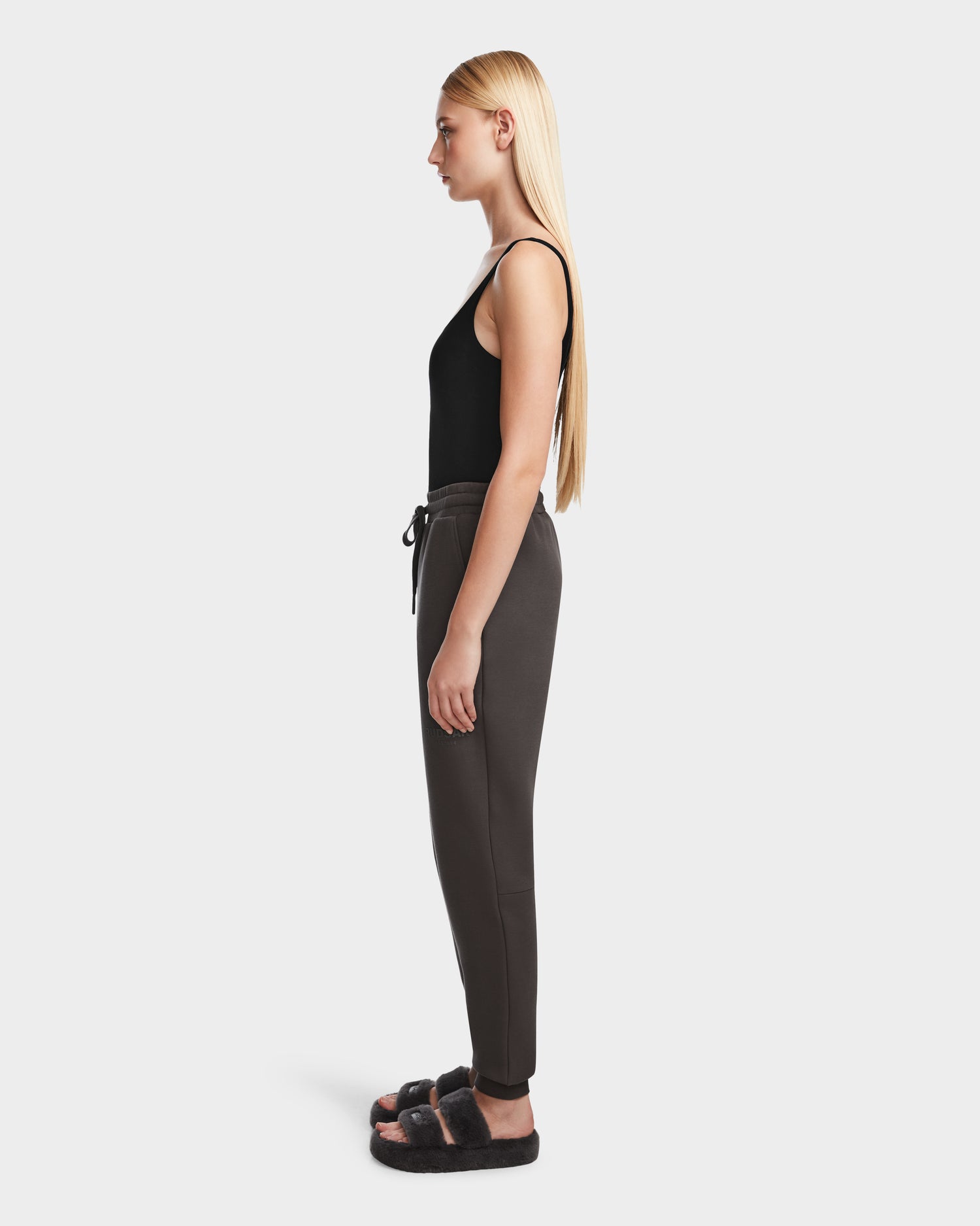 A woman with long blonde hair stands in profile, wearing Rudsaks JOSS - 9524009 Grey W premium cotton jogger pants and a black sleeveless top, paired with black fuzzy slide sandals against a plain white background.