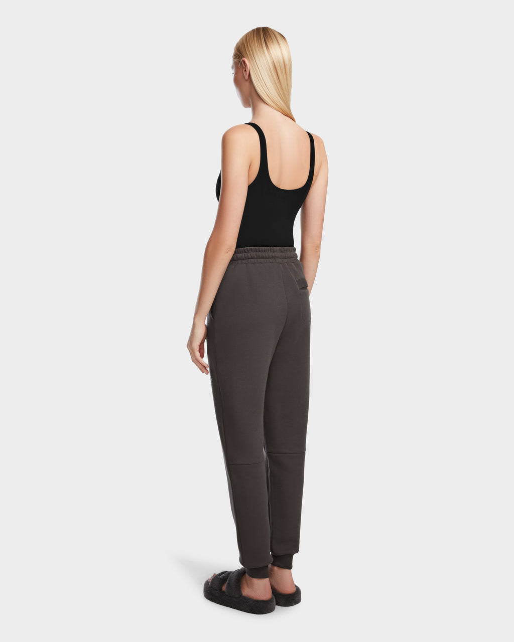 A woman with straight blonde hair stands facing away in Rudsak’s JOSS - 9524009 Grey W jogger sweatpants, a black tank top, and slide sandals against a plain white background.