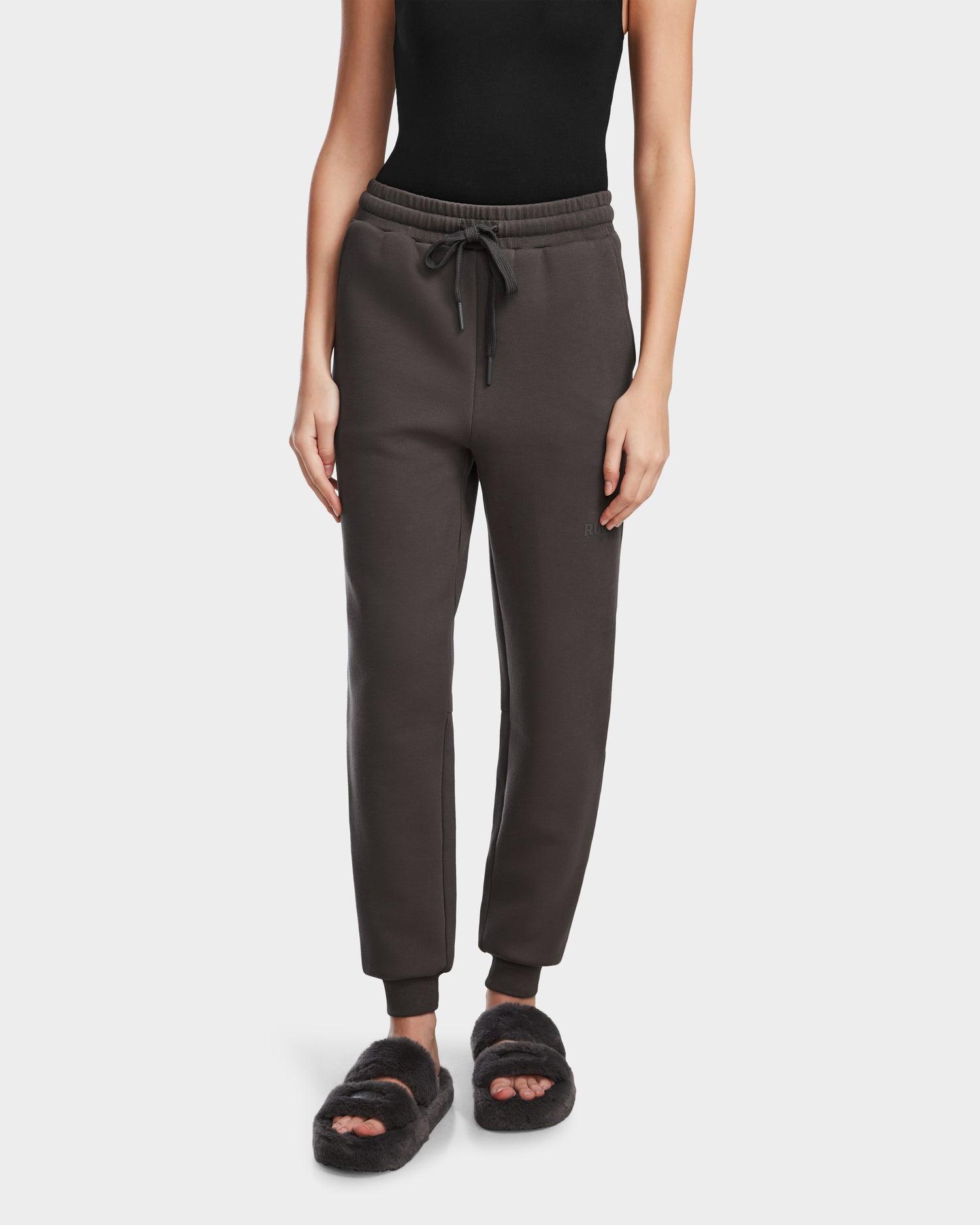 A person wears the Rudsak JOSS - 9524009 Grey W jogger sweatpants with a drawstring waist, paired with a black sleeveless top and black fuzzy open-toe slippers, standing against a plain white background.