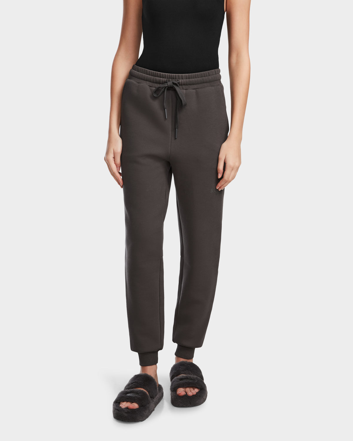 A person wears the Rudsak JOSS - 9524009 Grey W jogger sweatpants with a drawstring waist, paired with a black sleeveless top and black fuzzy open-toe slippers, standing against a plain white background.