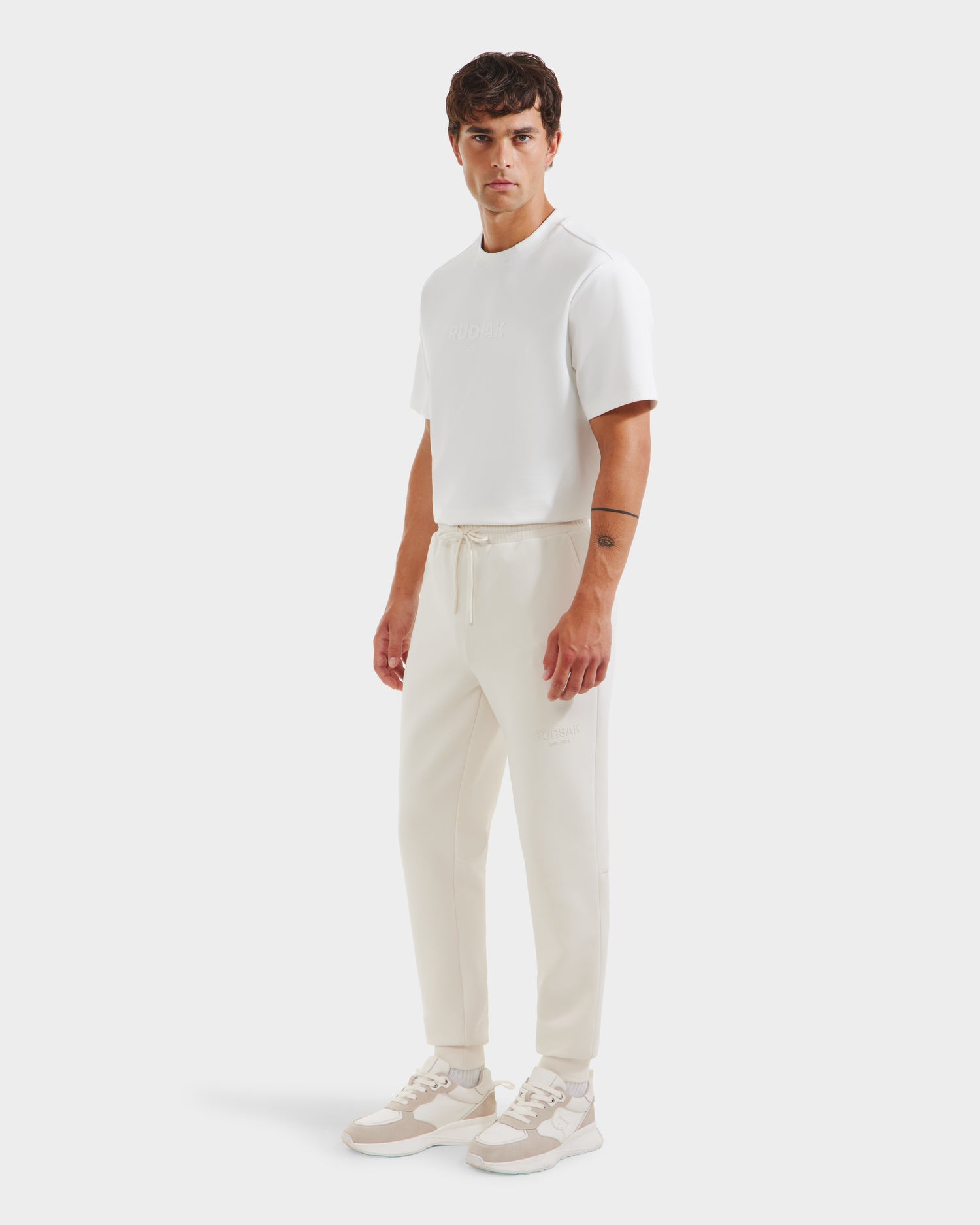 A man wearing the Rudsak JOSS - 9524009 Vanilla M casual outfit—premium cotton blend shirt and jogger sweatpants with white sneakers—stands against a plain white background.