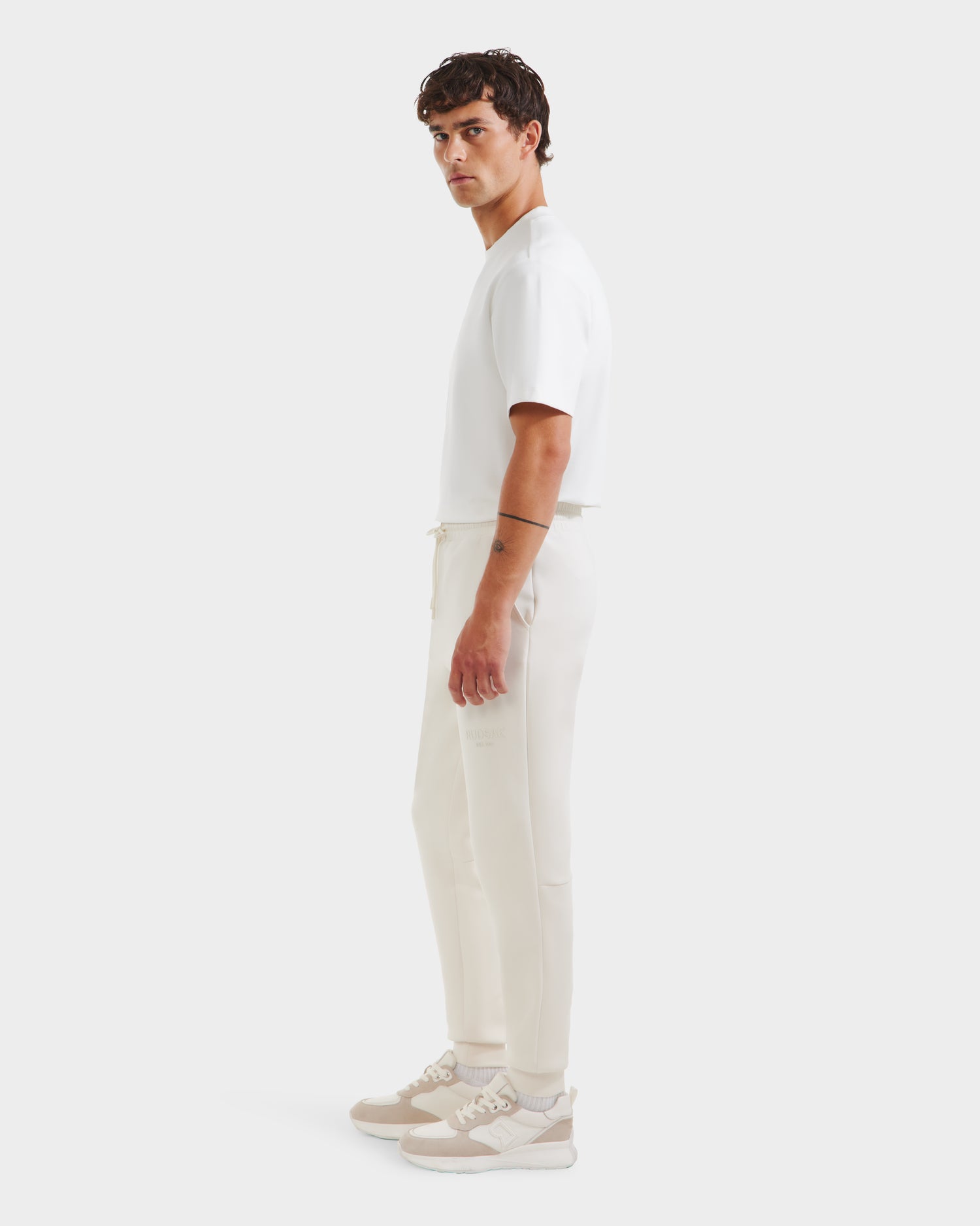 A man in profile against a white background wears Rudsak’s JOSS - 9524009 Vanilla M premium cotton blend off-white jogger pants, paired with a white T-shirt and beige sneakers. His curly dark hair adds to his clean, modern look.