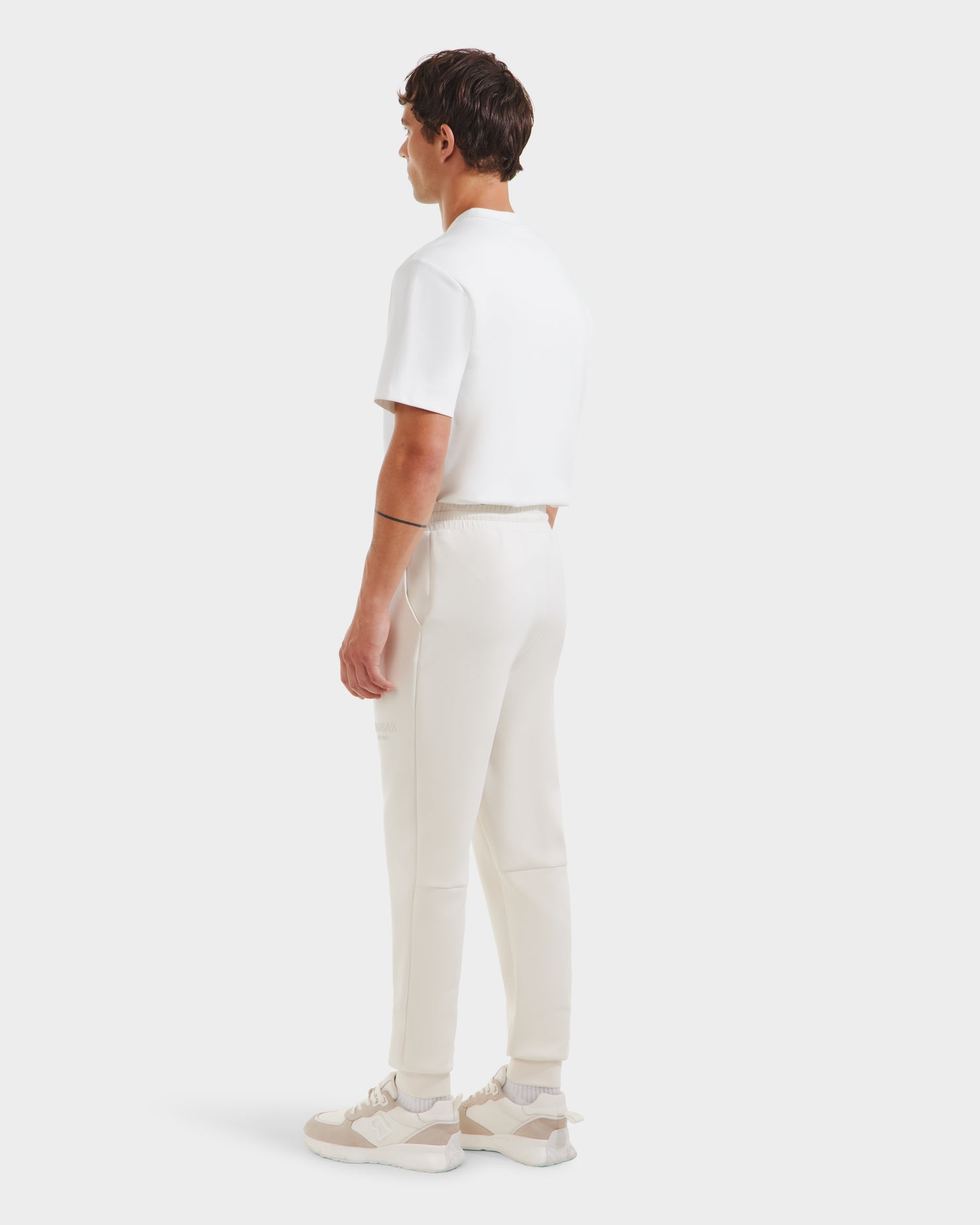 A man faces away, wearing the Rudsak JOSS - 9524009 Vanilla M premium cotton blend short-sleeve shirt, modern joggers, and white sneakers with beige accents, set against a plain white background.