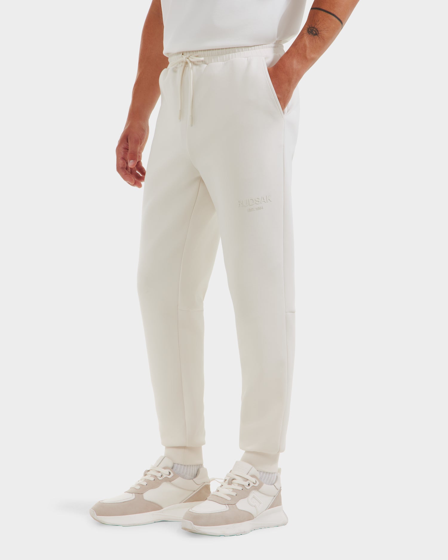 A person wears Rudsak’s JOSS - 9524009 Vanilla M unisex jogger pants with a drawstring waist, styled with matching sneakers and a white shirt, standing against a plain white background.