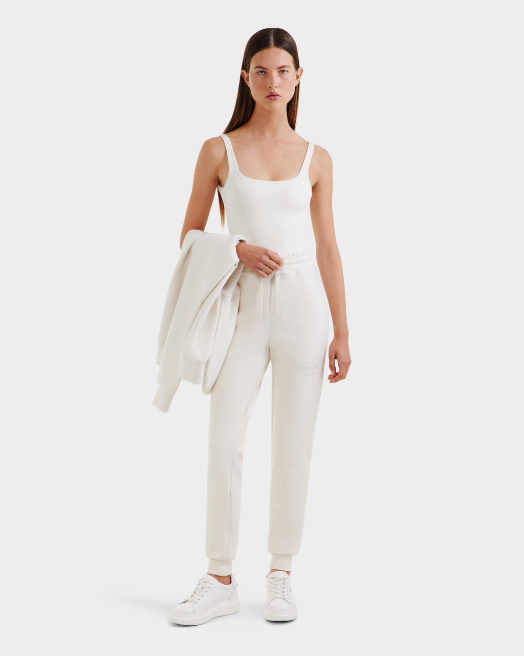 A woman stands against a plain background wearing the Rudsak JOSS - 9524009 Vanilla W joggers—a white tank top, premium cotton joggers, and white sneakers—while holding a matching white jacket over her arm.