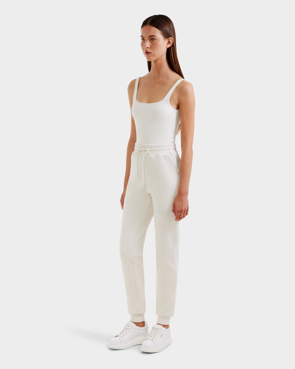 A woman with straight, dark hair and a neutral expression wears the Rudsak JOSS - 9524009 Vanilla W premium cotton joggers with a drawstring, styled with a fitted white tank top and white sneakers against a plain background.