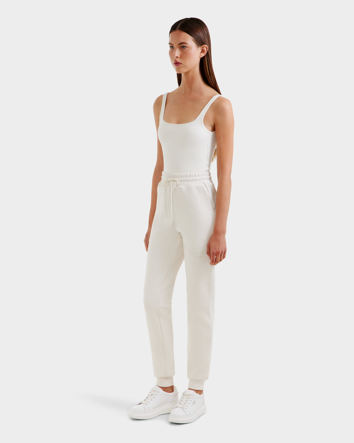 A woman with straight, dark hair and a neutral expression wears the Rudsak JOSS - 9524009 Vanilla W premium cotton joggers with a drawstring, styled with a fitted white tank top and white sneakers against a plain background.