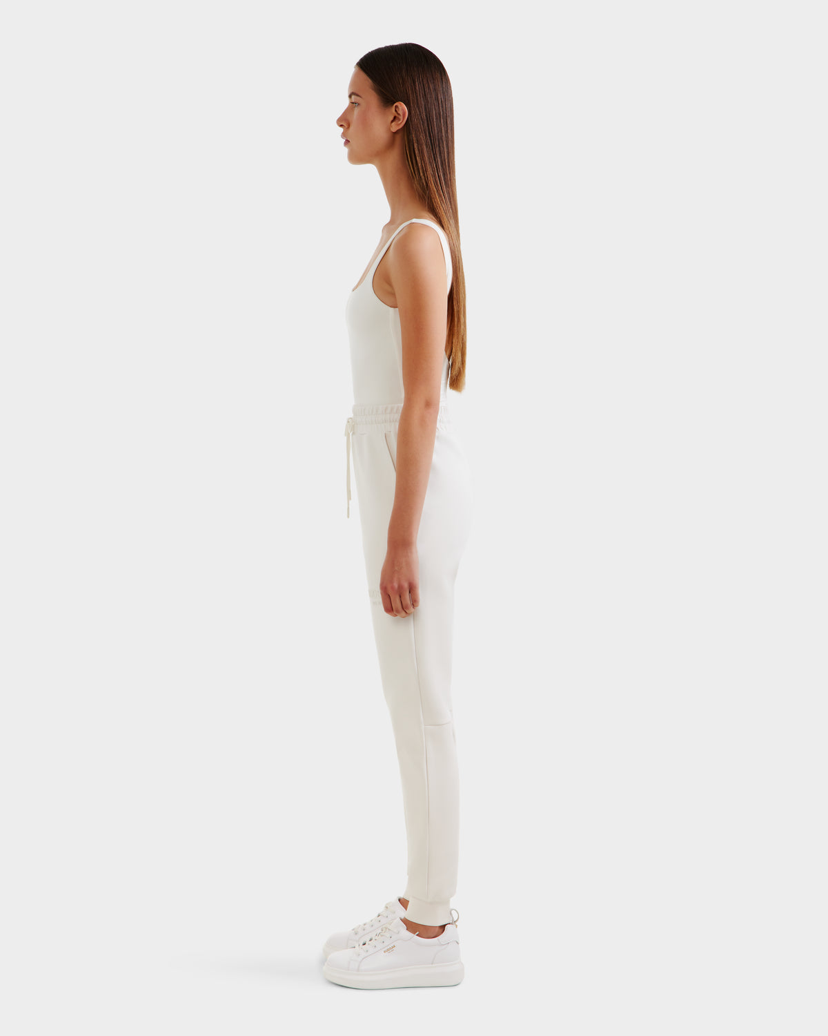 A woman with long brown hair stands in profile against a white background, wearing the Rudsak JOSS - 9524009 Vanilla W joggers, a sleeveless white top, and white sneakers, with her arms resting naturally at her sides.