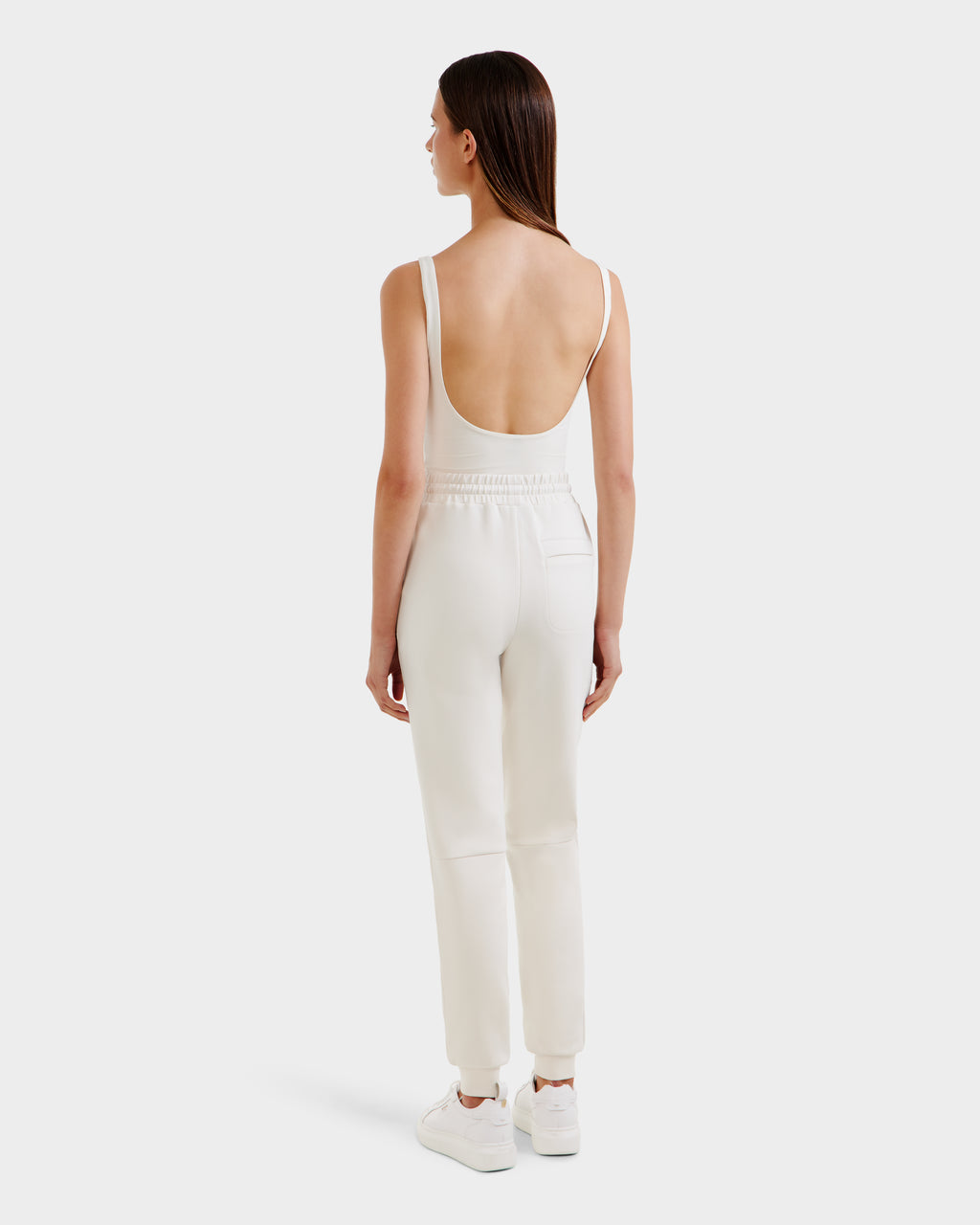 A woman with straight brown hair models the Rudsak JOSS - 9524009 Vanilla W sleeveless white jumpsuit with a low back, styled with joggers and white sneakers, standing against a plain white background, facing away.