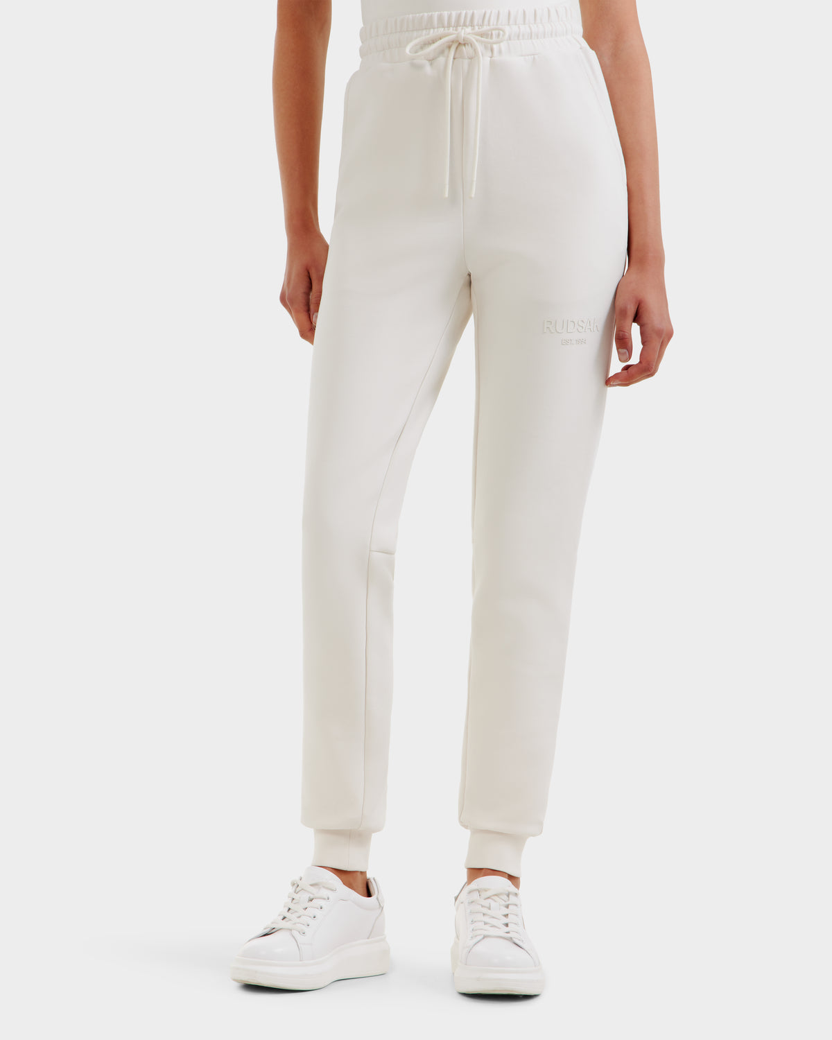 A person wears Rudsak's JOSS - 9524009 Vanilla W premium cotton joggers in off-white, featuring a drawstring waist and paired with white sneakers, shown from waist to feet against a white background.