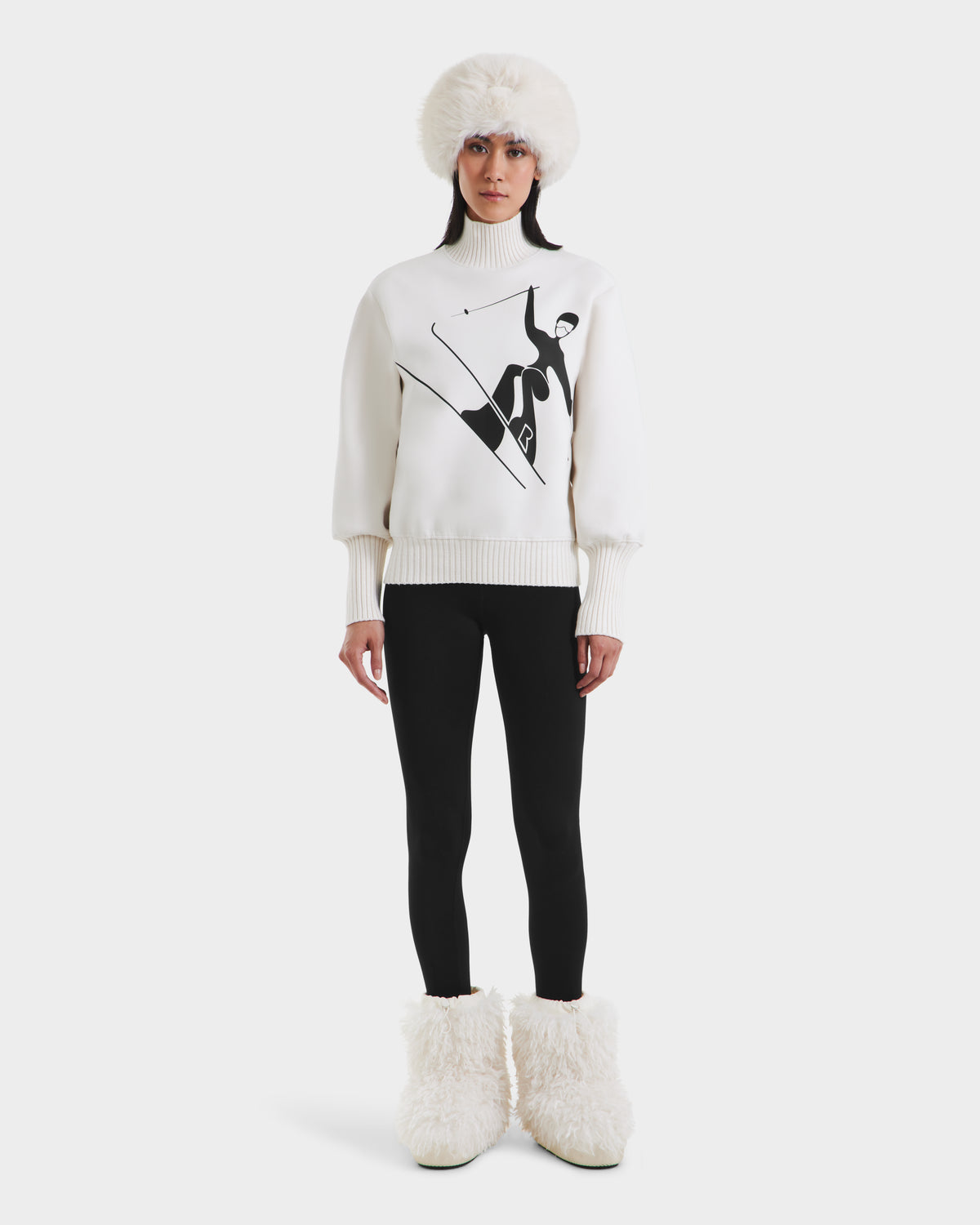 A woman faces forward against a plain light background, wearing the Rudsak JOYCE - 8525647 Vanilla ski sweater with a black skiing graphic, paired with a white fur hat, black leggings, and large white furry boots.