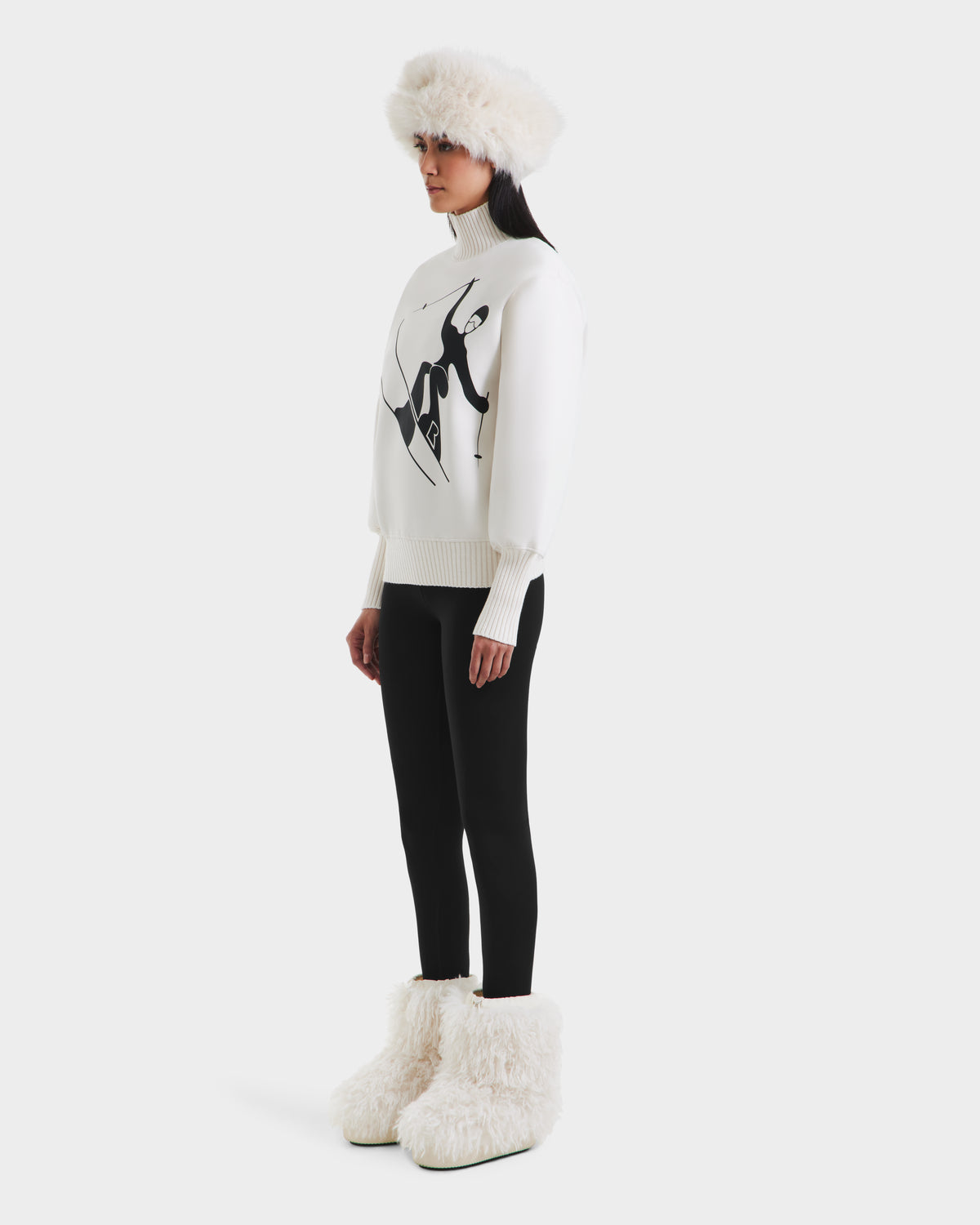 A woman stands sideways in the Rudsak JOYCE - 8525647 Vanilla cozy cotton-blend women’s ski sweater with a black abstract design, paired with a white furry hat, black leggings, and large white furry boots—perfect après-ski style.
