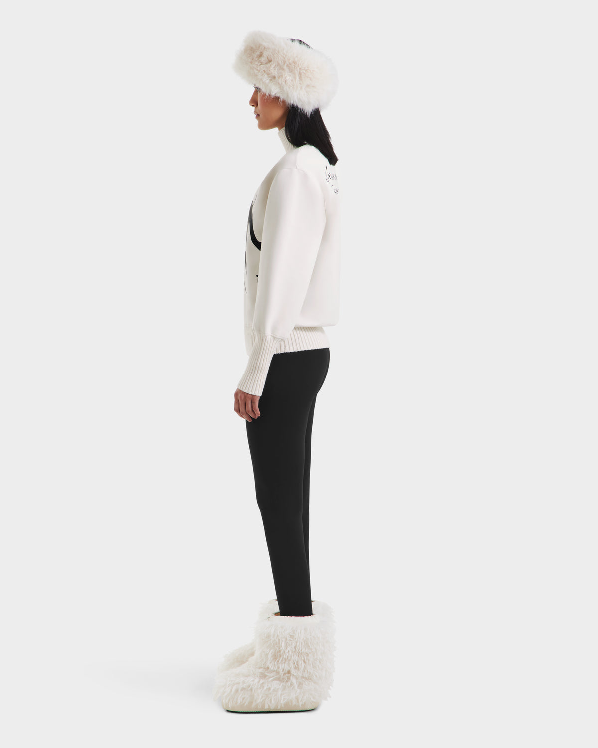 A woman in profile wears the JOYCE - 8525647 Vanilla jacket by Rudsak with a white fur hat, après-ski sweater, black leggings, and oversized white furry boots against a plain white background.