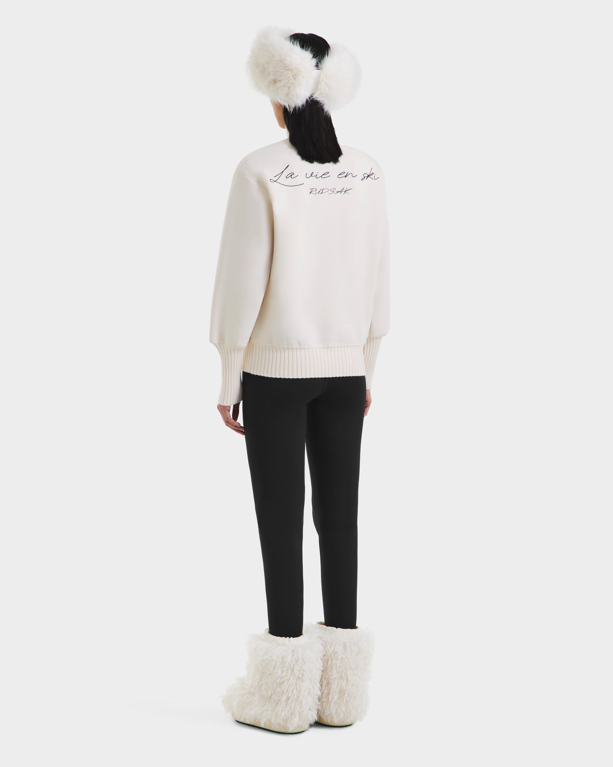 A person stands facing away, wearing the Rudsak JOYCE - 8525647 Vanilla retro-inspired ski sweater with text on the back, paired with black pants, fluffy white earmuffs, and large furry white boots against a plain white background.