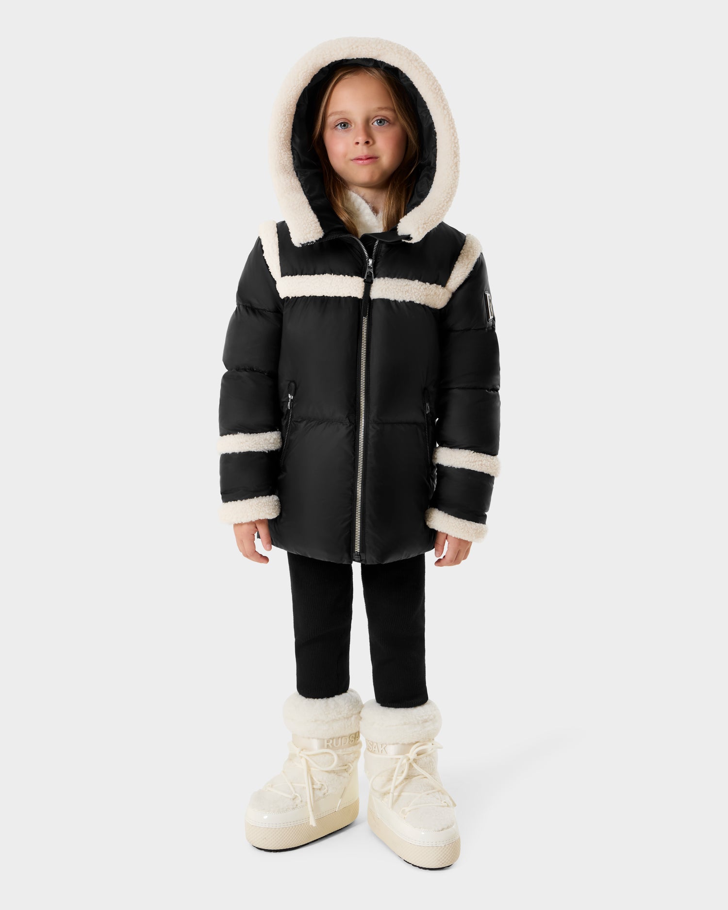 A young child wears the Rudsak JULIANA K - K125550 Black puffer jacket with white fur trim and hood, paired with black pants and white fur-lined boots, standing against a plain light gray background.