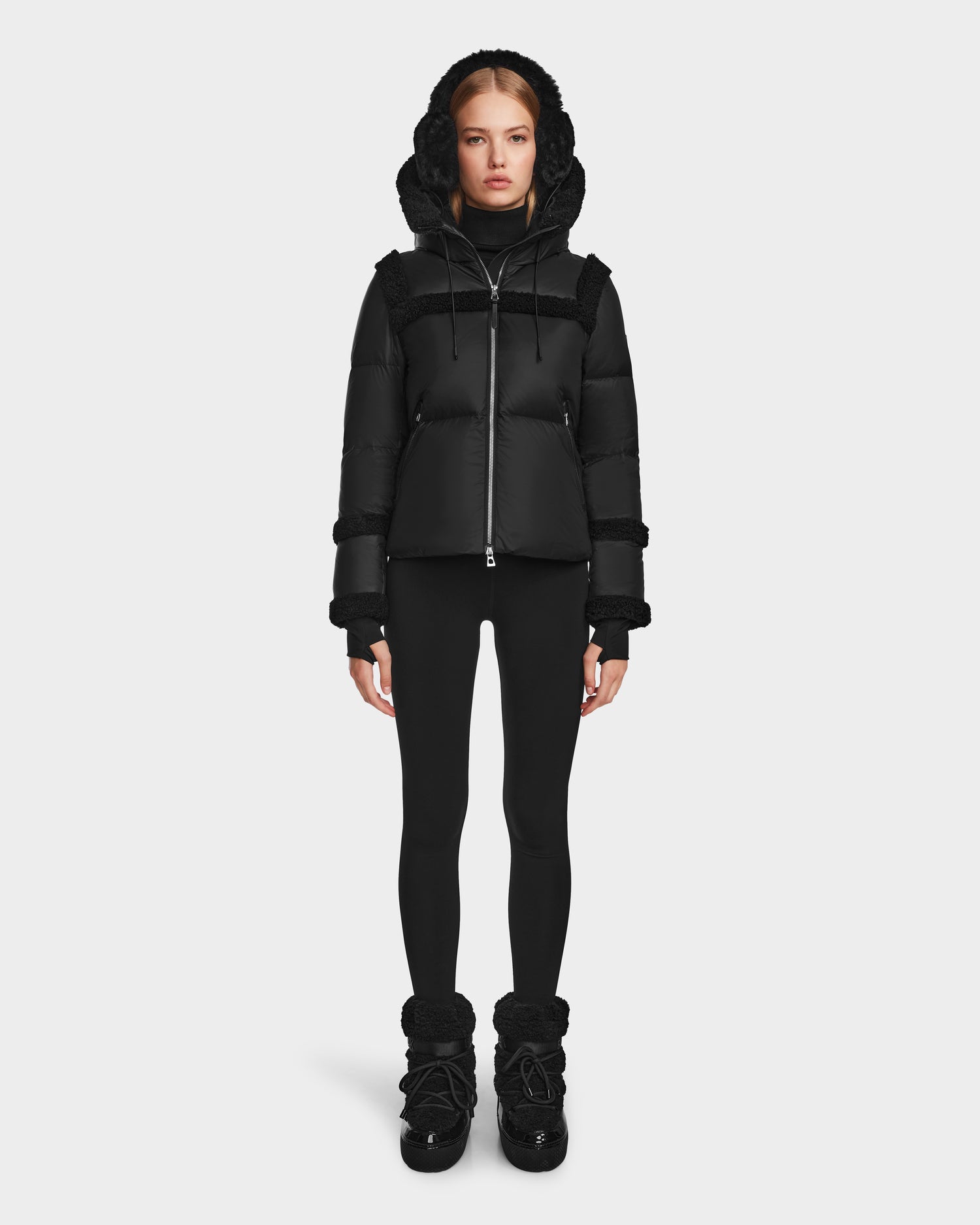 A woman stands facing forward against a plain white background wearing the Rudsak JULIANA - L124509 Black/Black women's short down puffer with a hood, black leggings, and black winter boots.
